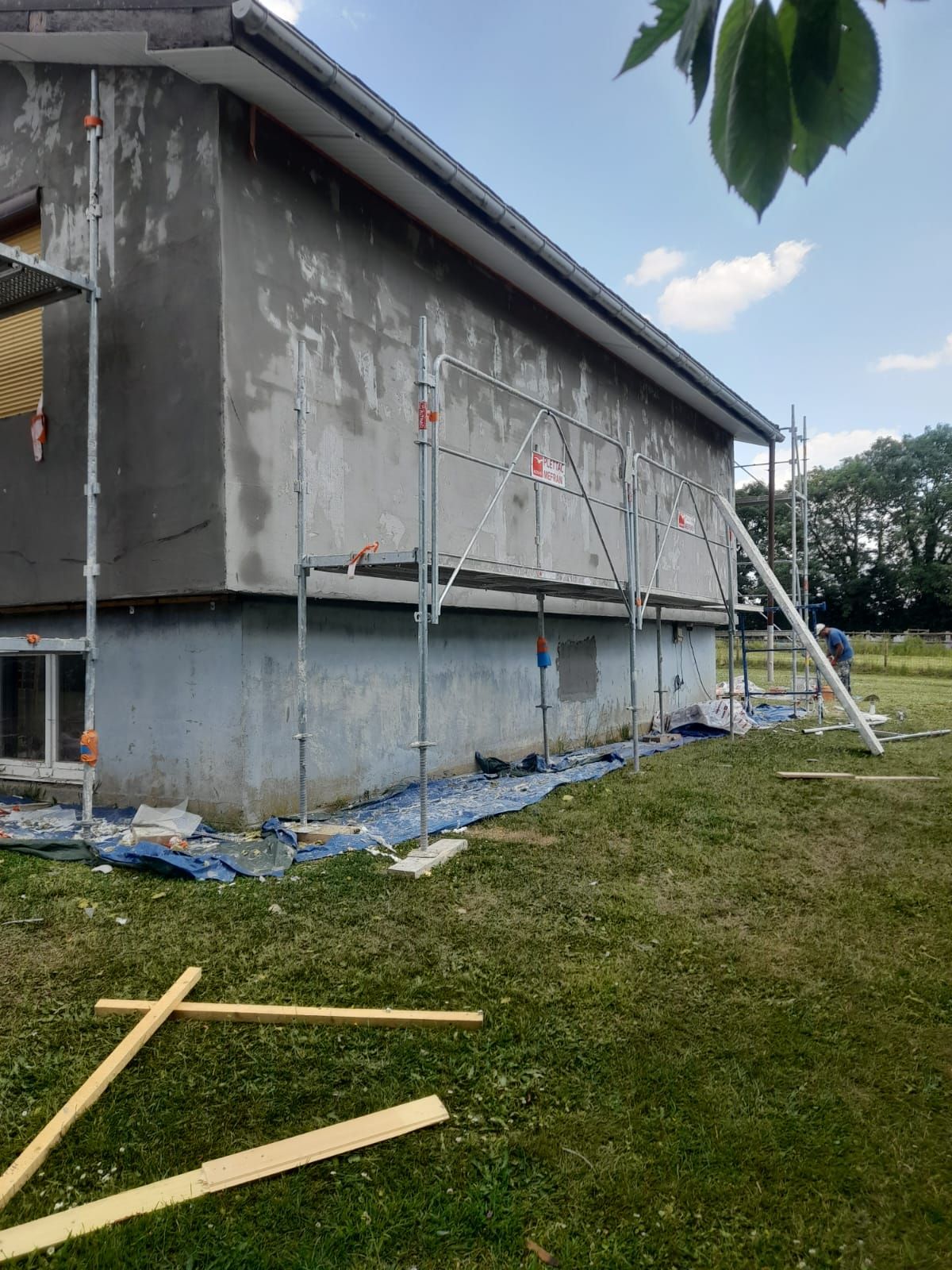 Façade d'un bâtiment en construction avec échafaudages. Revêtement en stuc gris, bâche bleue, pelouse verte, ciel dégagé.