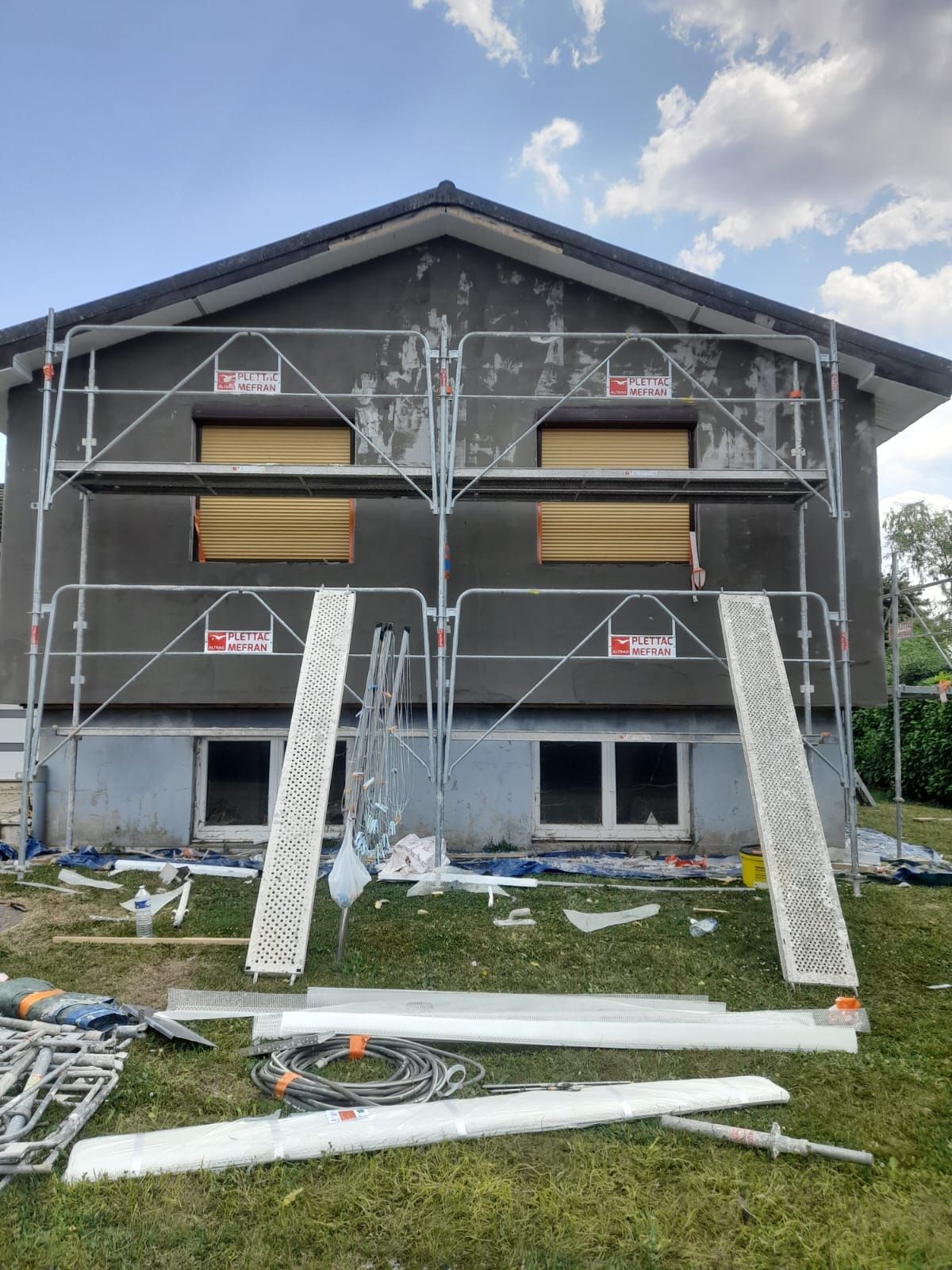 Façade d'une maison en rénovation, avec des échafaudages. Enduit gris, fenêtres condamnées et matériaux de construction au sol.