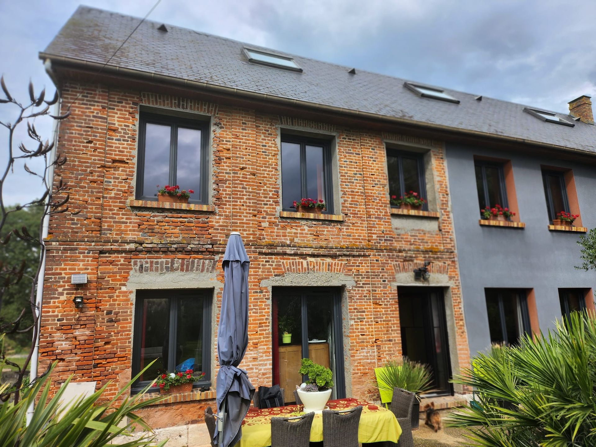 Maison en briques aux fenêtres sombres, toit gris et parasol bleu sur une terrasse avec des plantes.