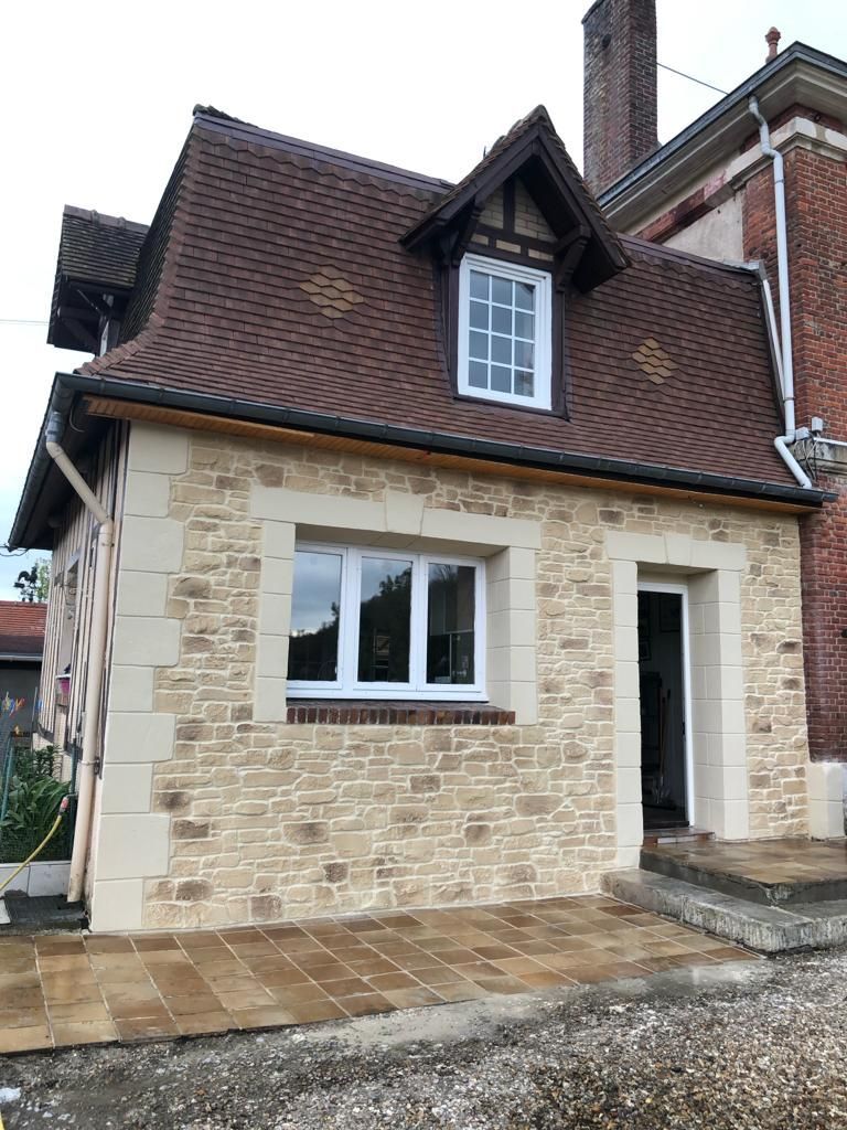 Petit bâtiment en pierre avec toit brun, fenêtres à encadrement blanc, détails en brique et porte ouverte.