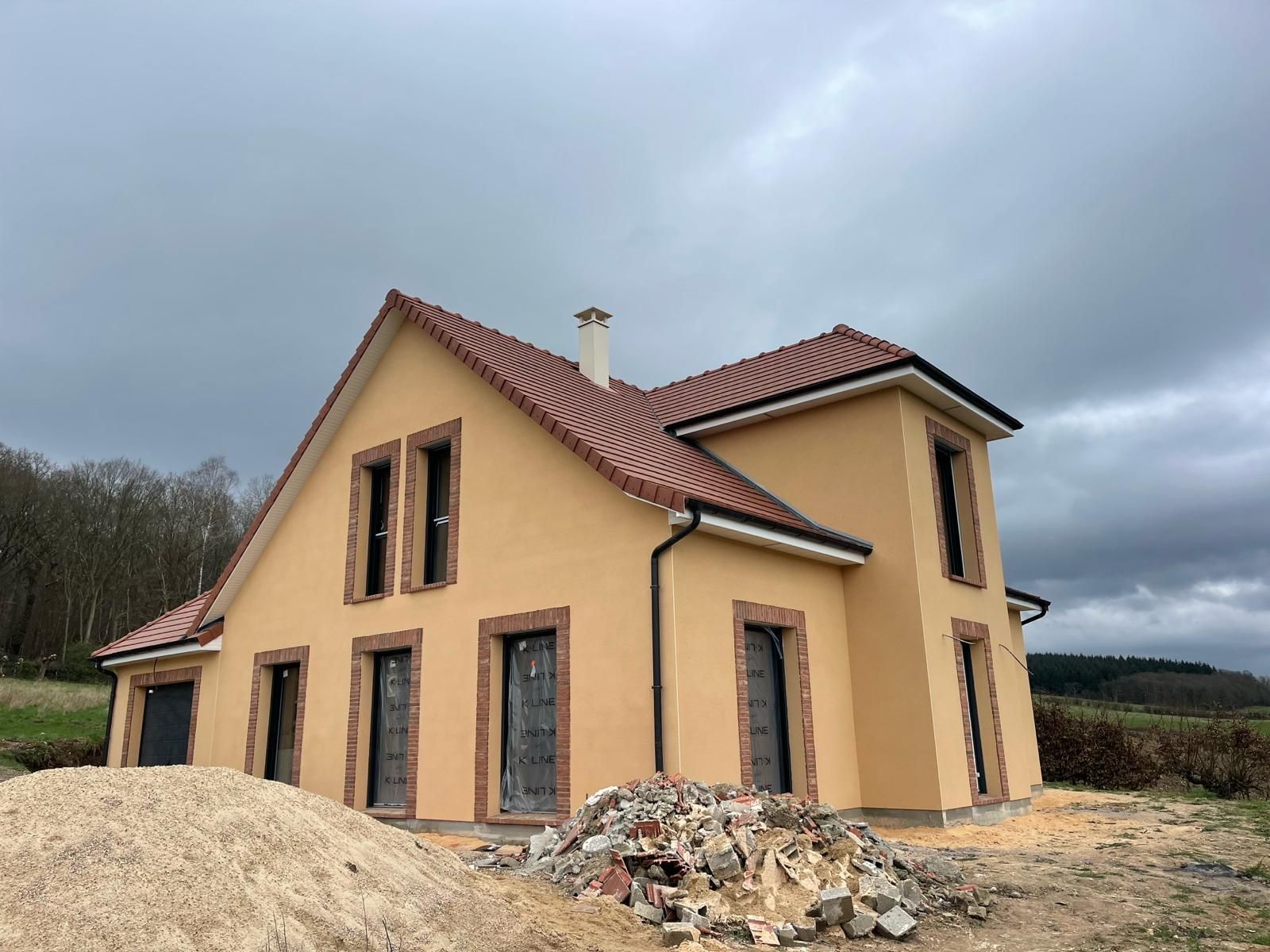 Maison beige en construction, avec un toit de tuiles rouges et un ciel gris foncé.