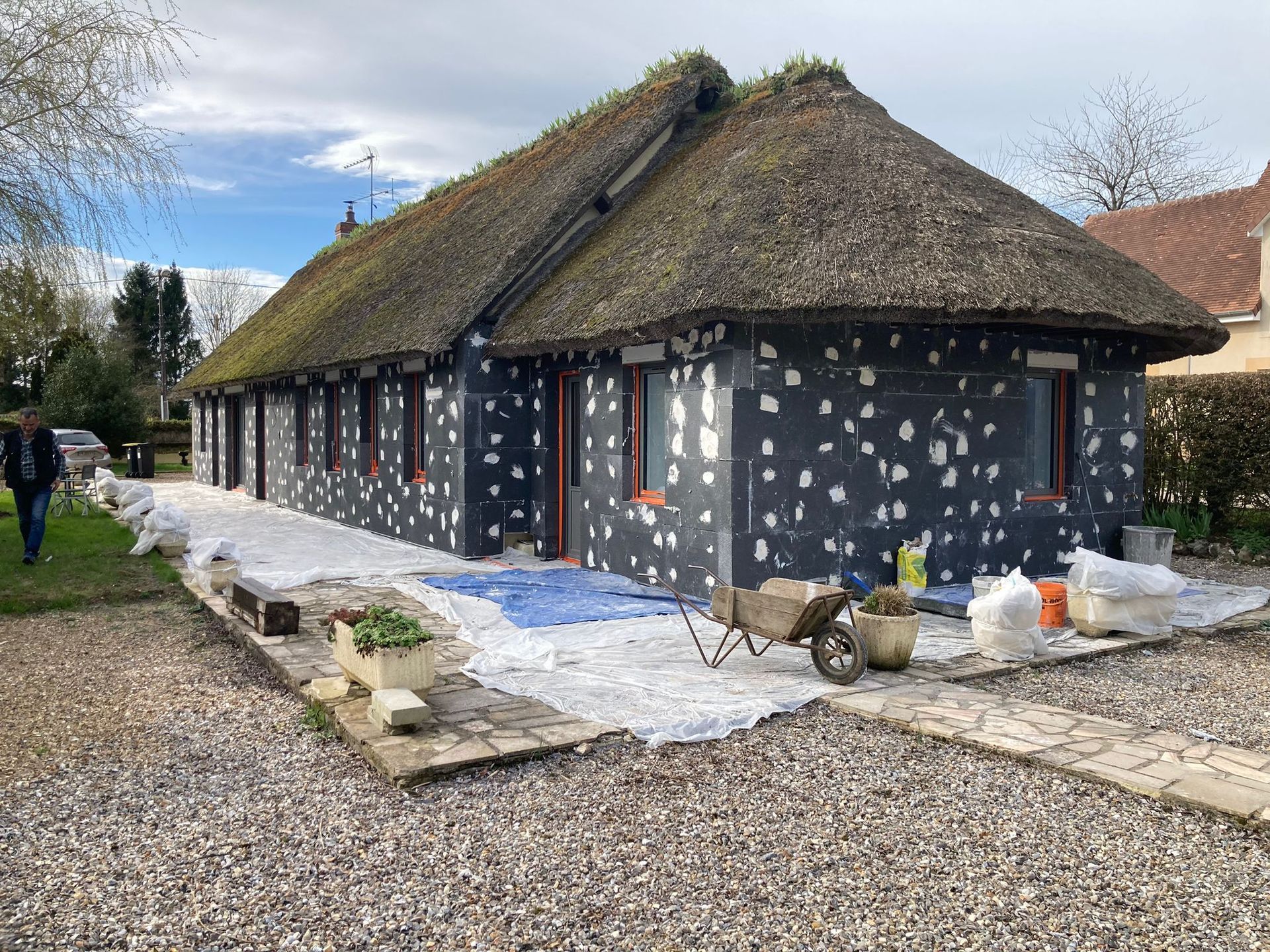 Maison au toit de chaume, façade noire tachetée de blanc, en cours de construction. Allée en gravier.
