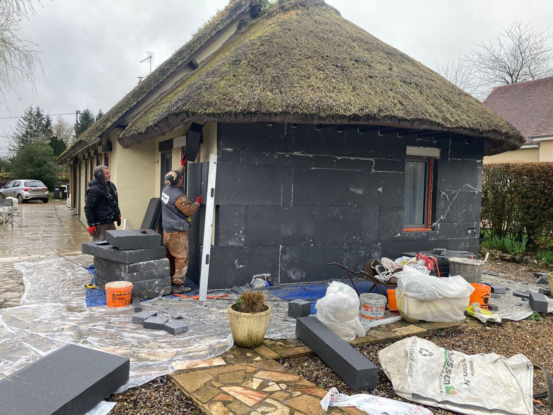 Des ouvriers installent de l'isolant noir sur le côté d'une maison au toit de chaume. Outils et matériaux sont éparpillés à proximité.