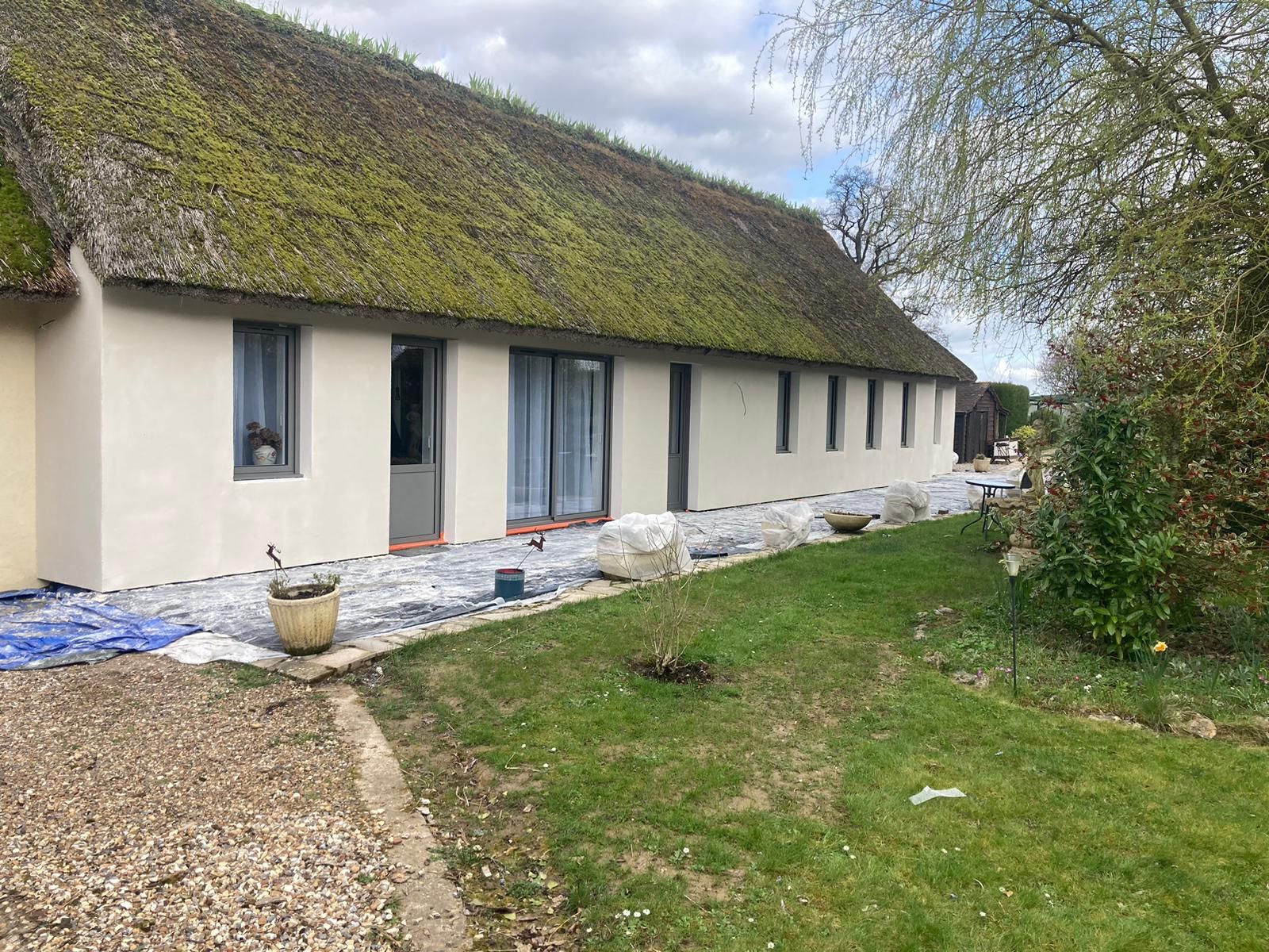 Maison beige au toit de chaume recouvert de mousse. Fenêtres et portes sur le côté. Allée de gravier et pelouse devant.