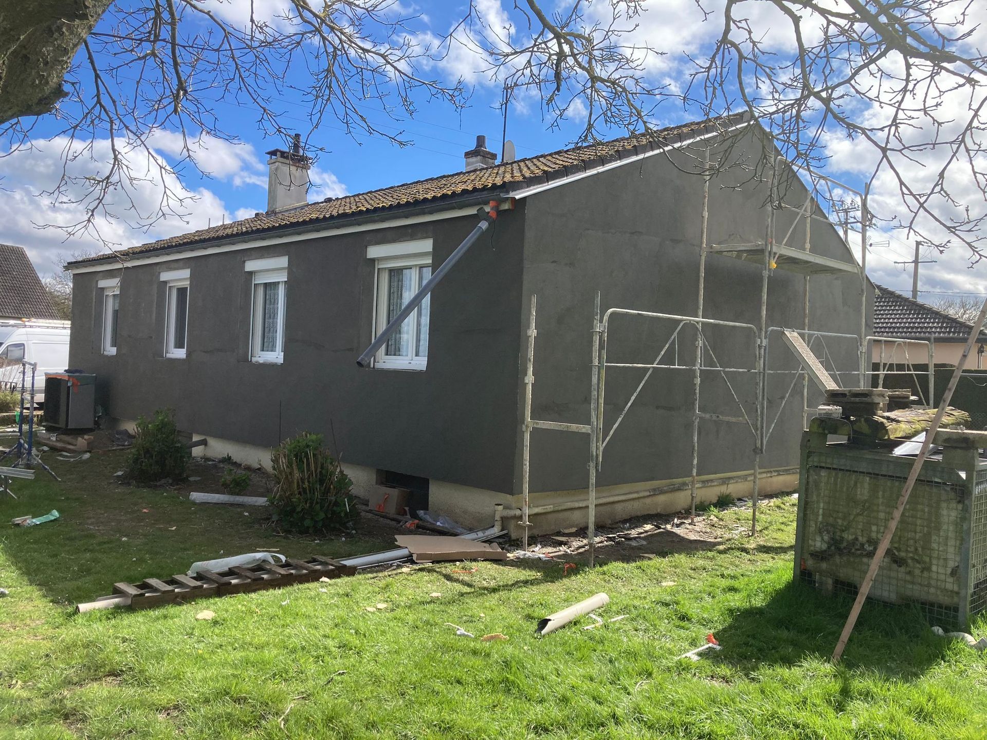 Maison grise avec des échafaudages, en cours de rénovation dans une cour verdoyante, sous un ciel bleu.