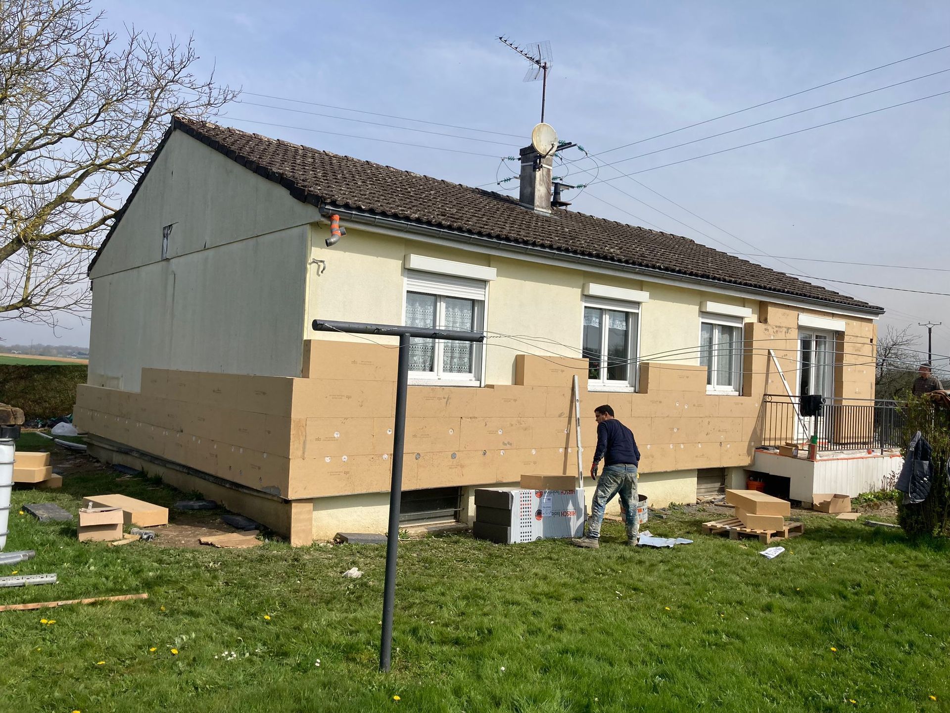 Un homme installe de l'isolant sur la façade d'une maison ; panneaux isolants beiges, bardage jaune, ciel bleu.