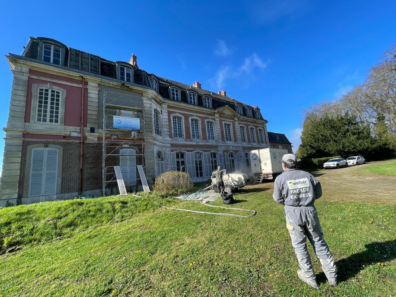 Un homme en combinaison grise observe un manoir historique en rénovation sur une colline herbeuse ; ciel bleu.