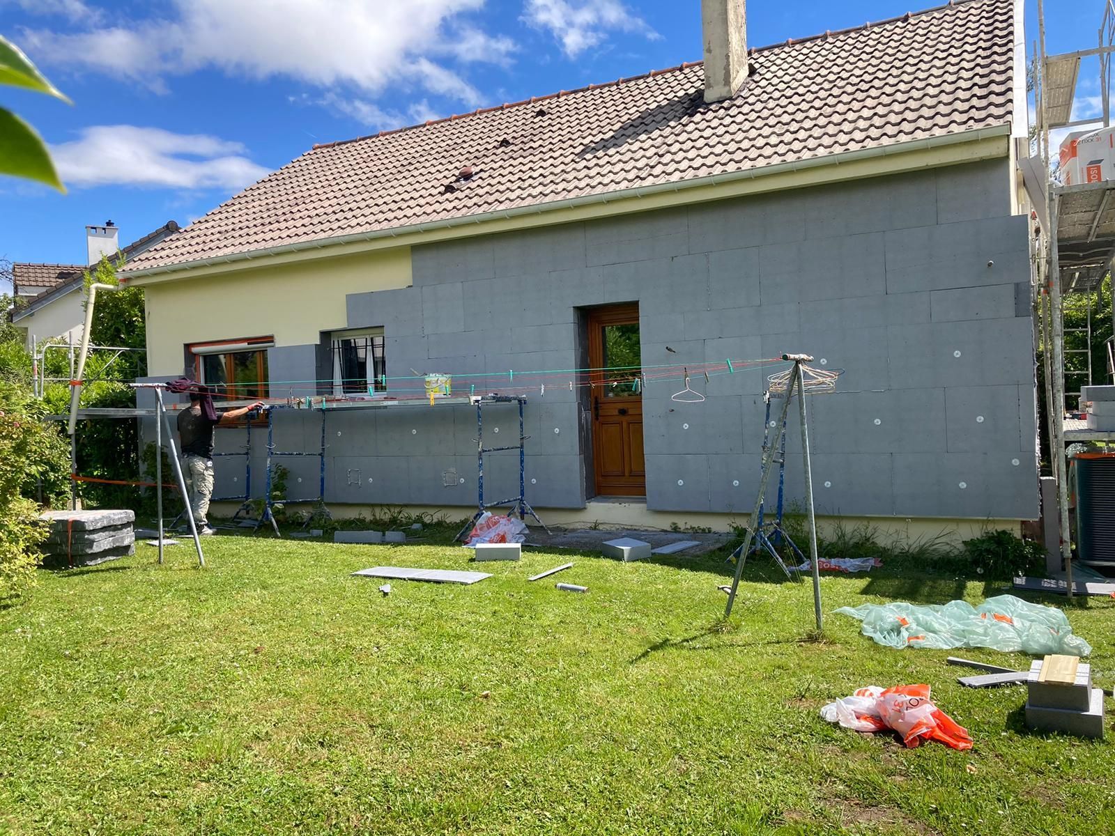 Façade d'une maison en cours d'isolation ; ouvrier sur un échafaudage. Journée ensoleillée, jardin verdoyant.