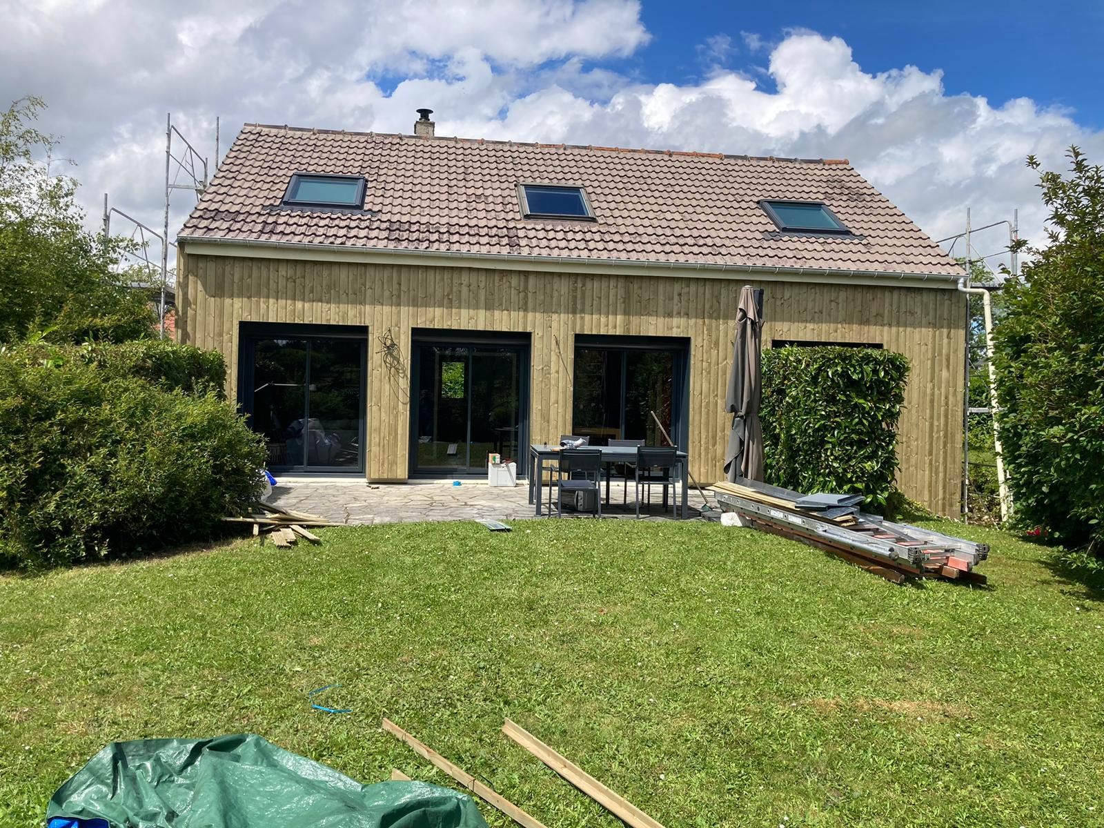 Maison en construction avec toiture apparente et bardage en bois. Grandes fenêtres et pelouse verte à l'avant.