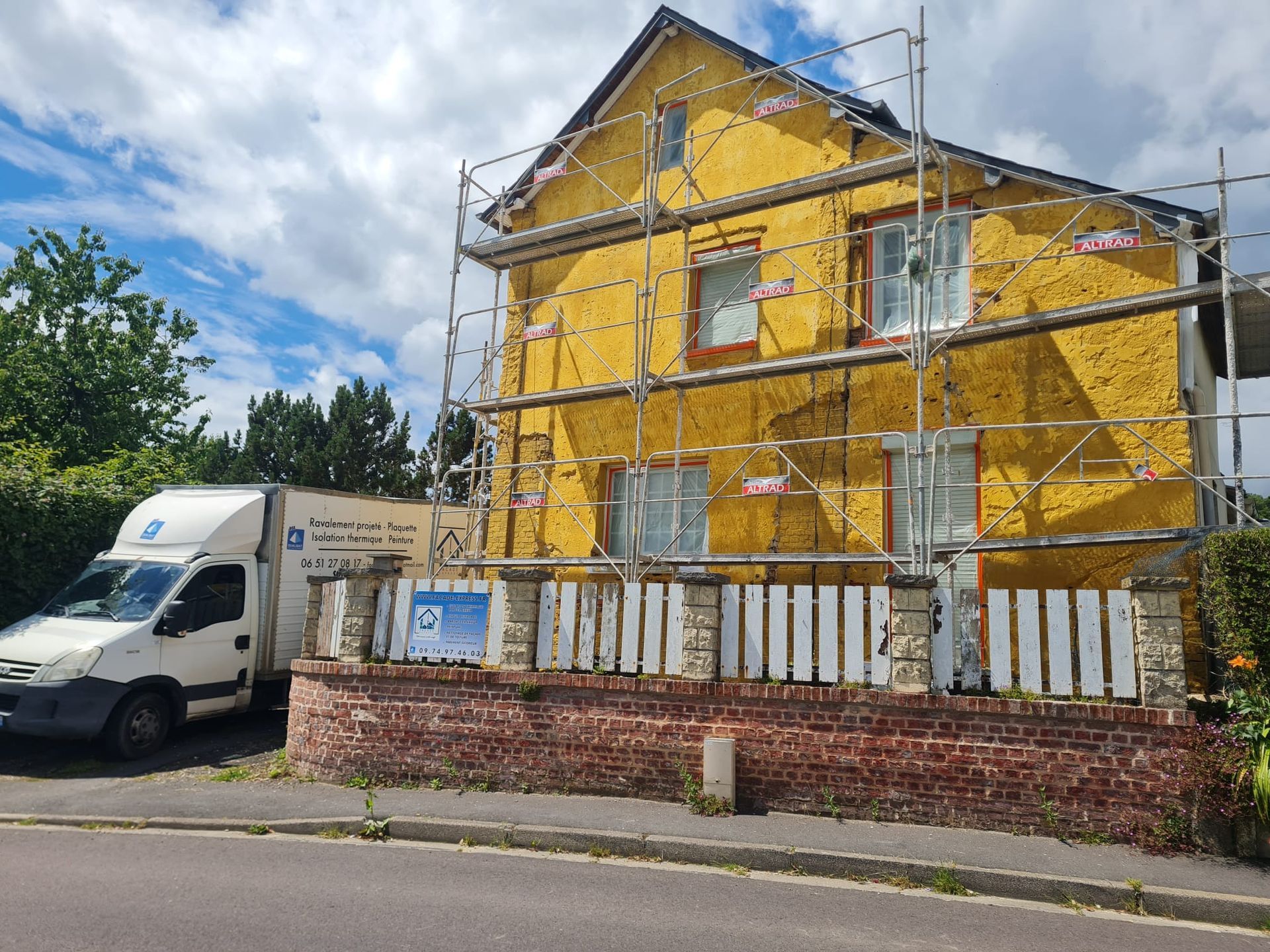 Maison jaune en construction avec des échafaudages, camion blanc garé devant.