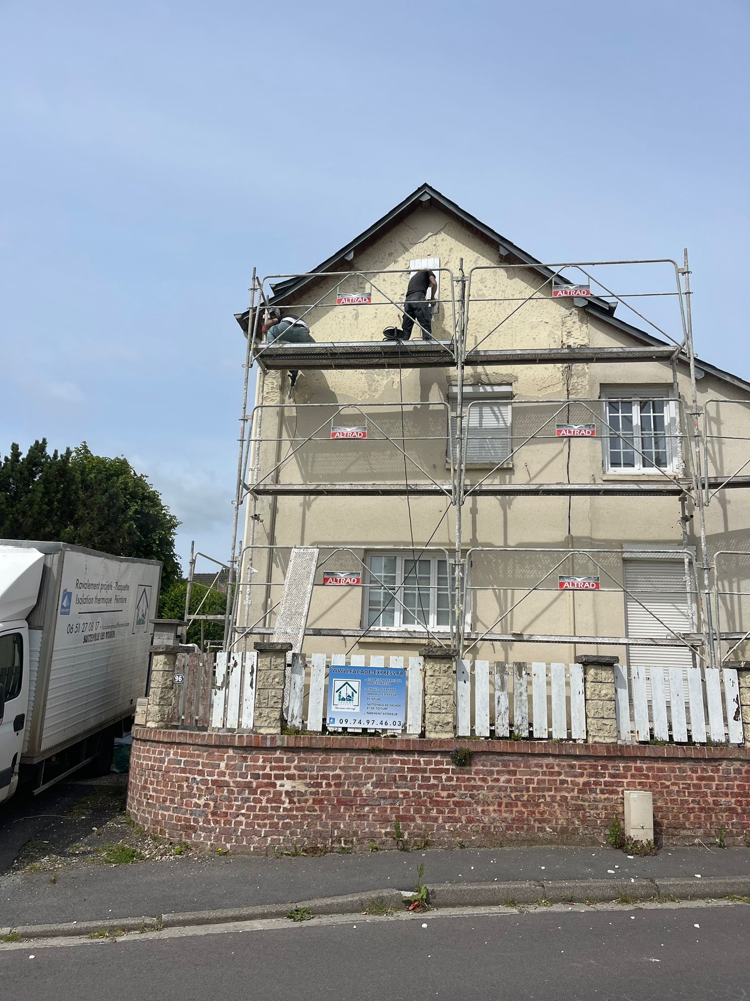 Façade du bâtiment avec échafaudages ; un ouvrier applique du crépi. Un camion est stationné à gauche.