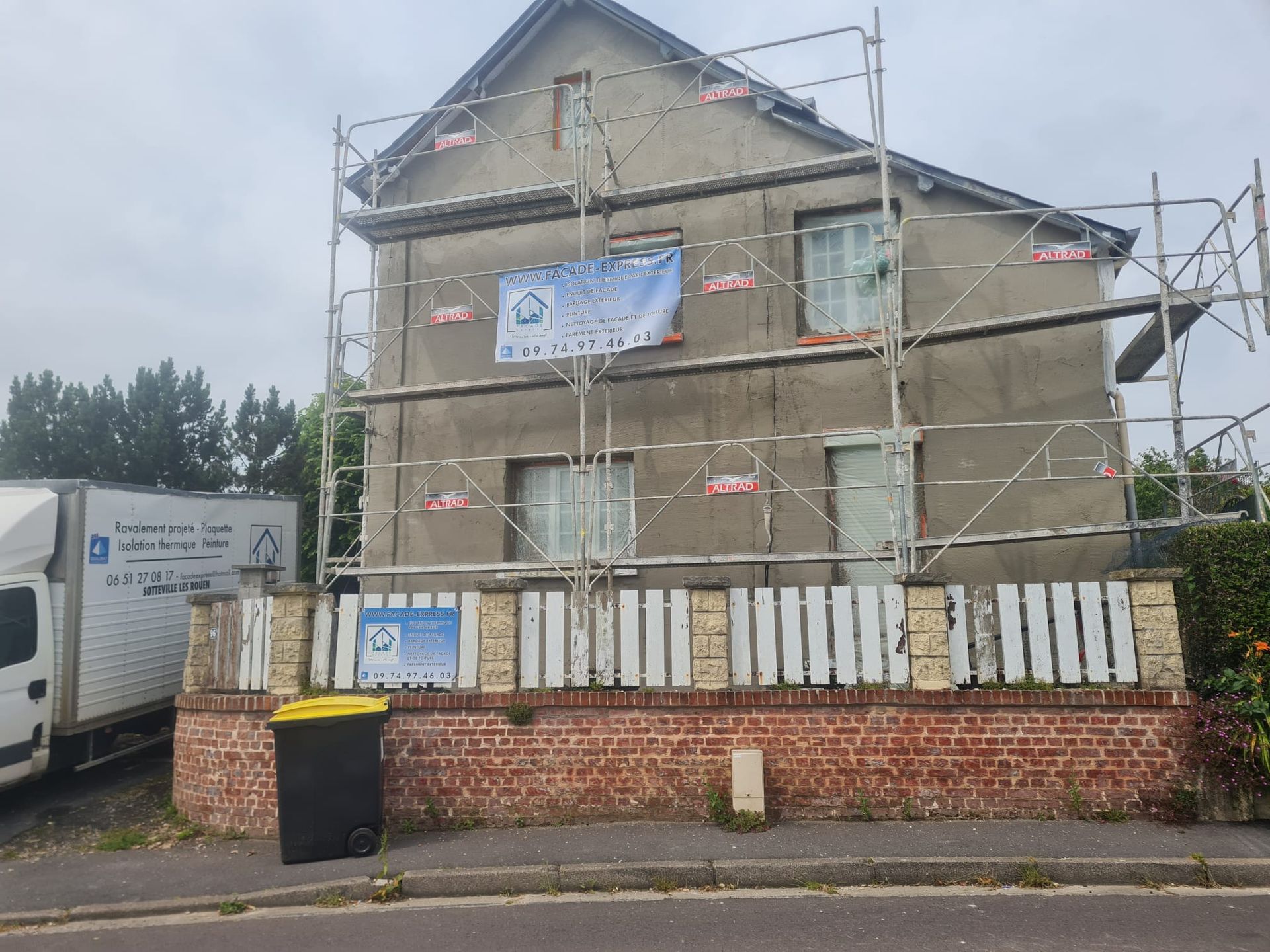 Extérieur de la maison en construction, échafaudages, façade grise, brique et clôture blanche, ciel couvert.