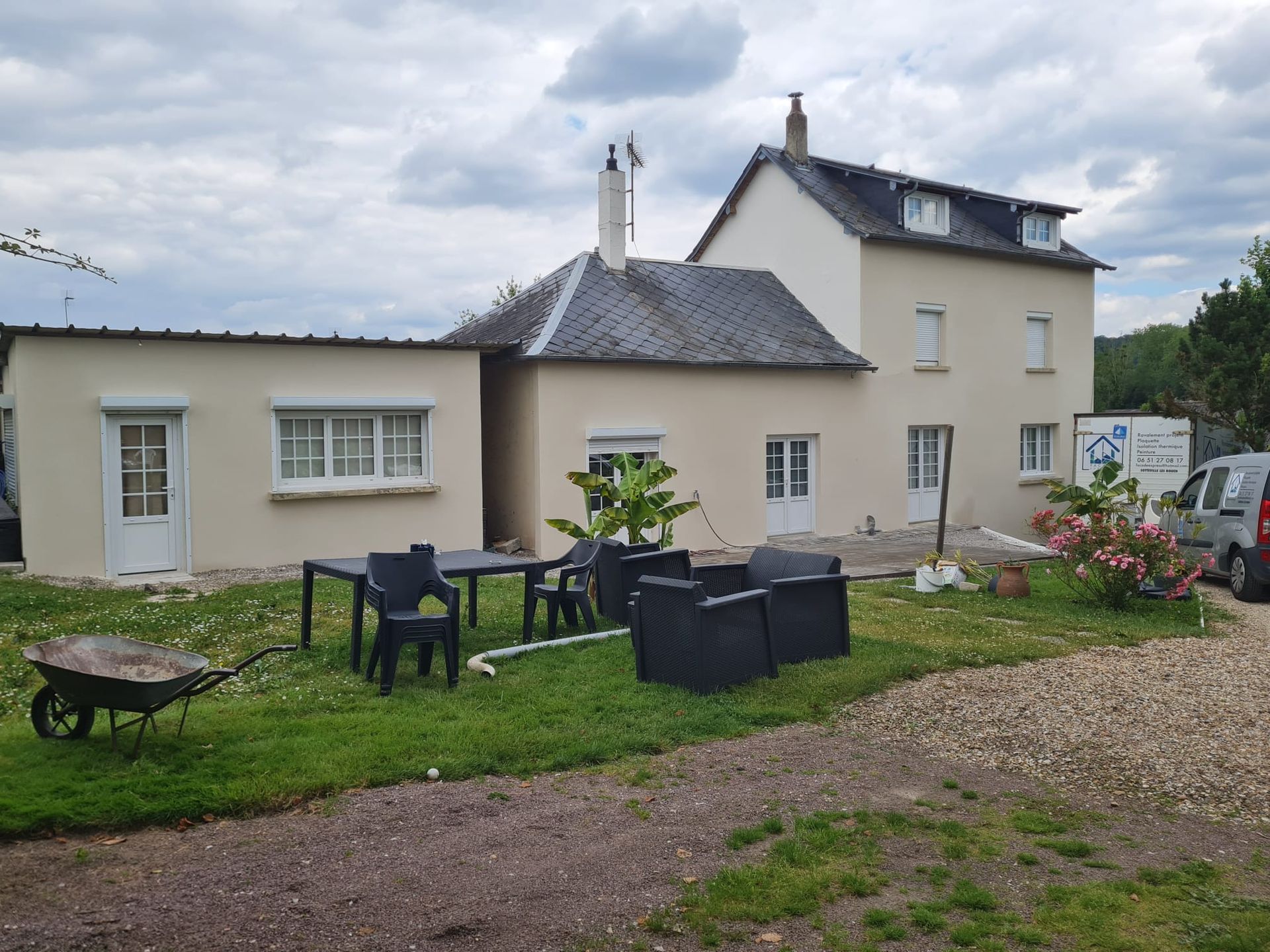 Une maison claire avec un jardin. Des meubles de jardin sont installés sur la pelouse. Une brouette se trouve au premier plan.