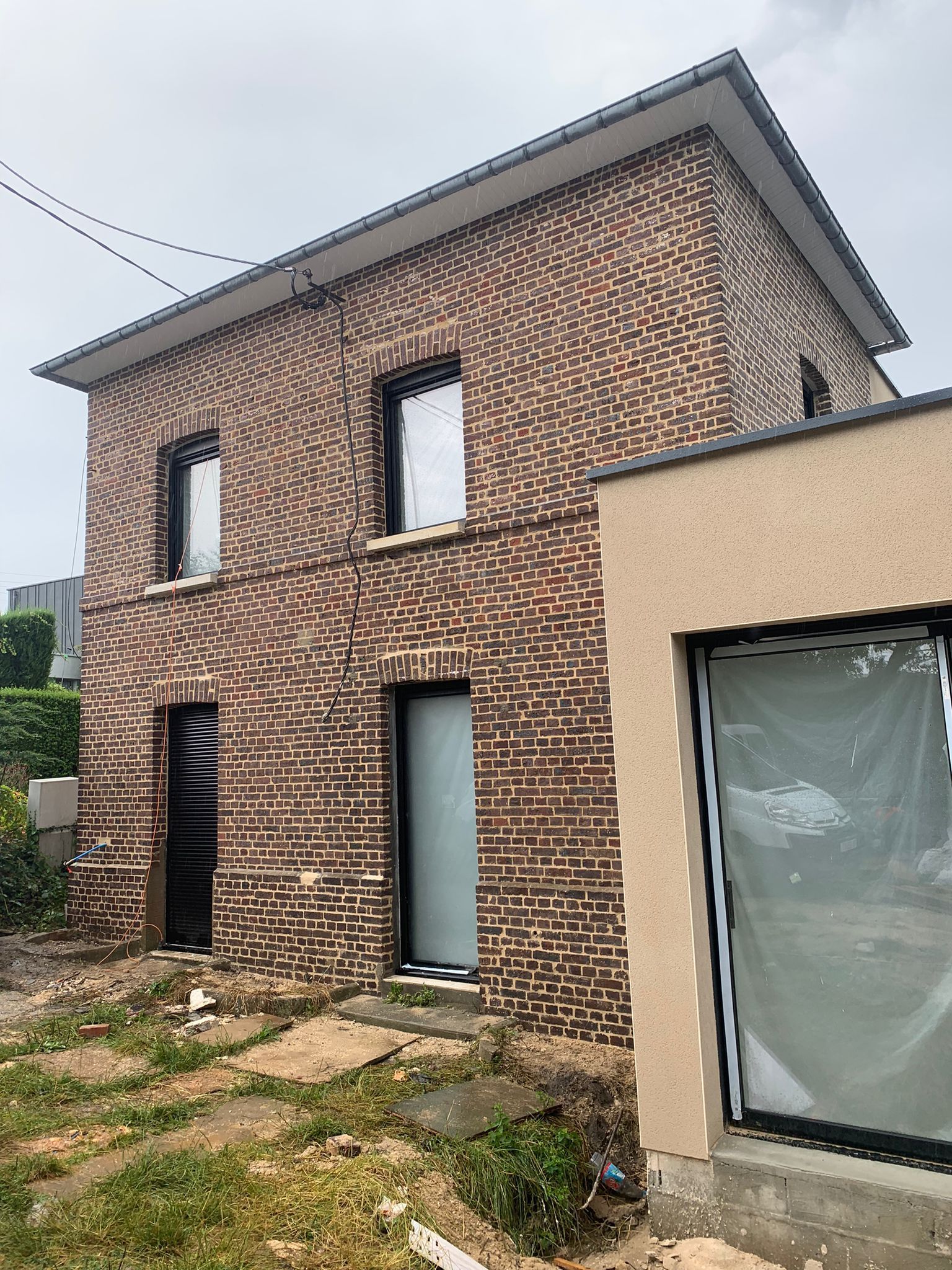Maison en briques en construction, avec fenêtres et portes à encadrement noir. Bâtiment attenant de couleur beige. Ciel couvert.