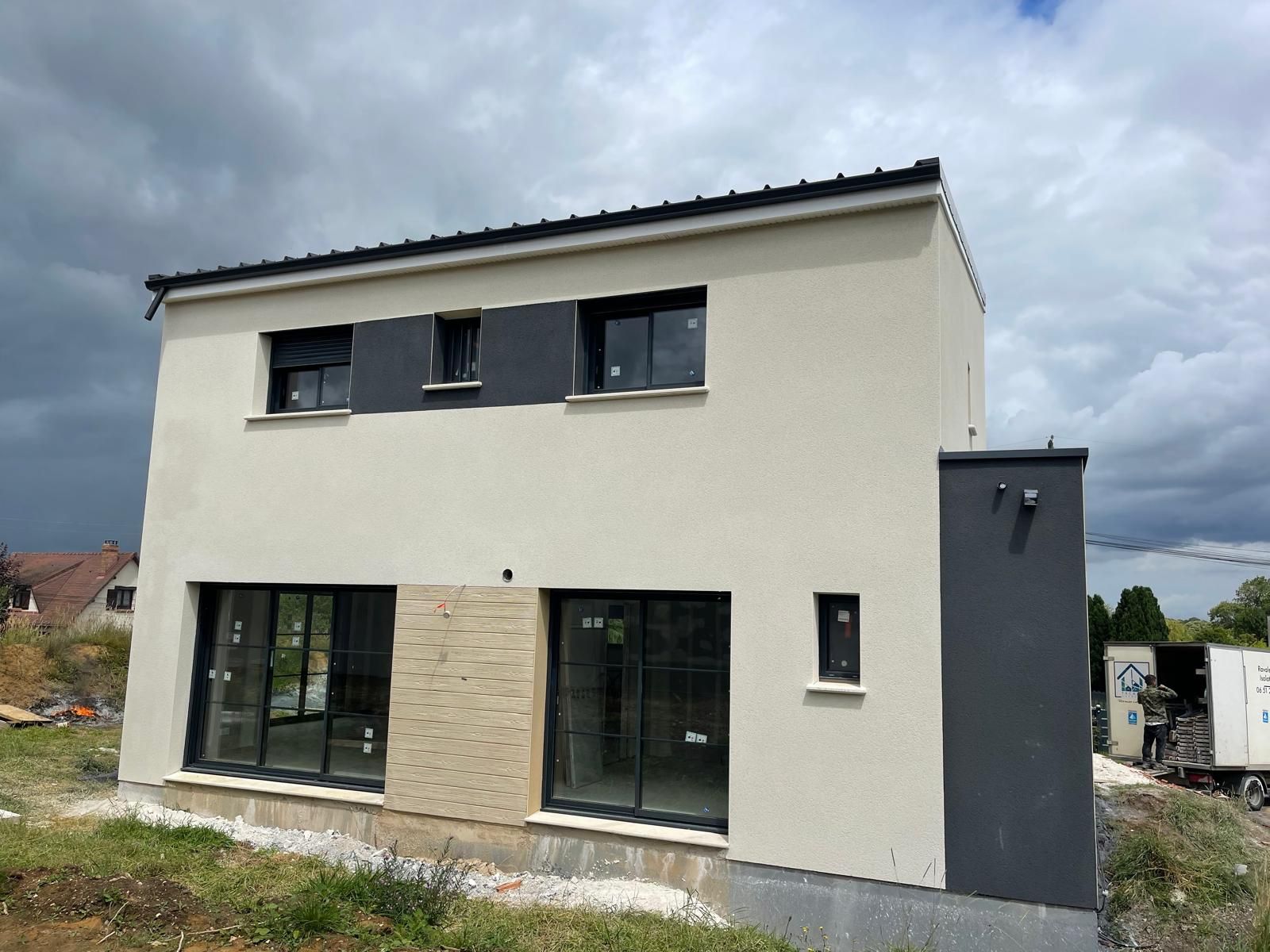 Maison à deux étages en construction, avec des murs extérieurs gris clair et gris foncé, de grandes fenêtres et un ciel nuageux en arrière-plan.