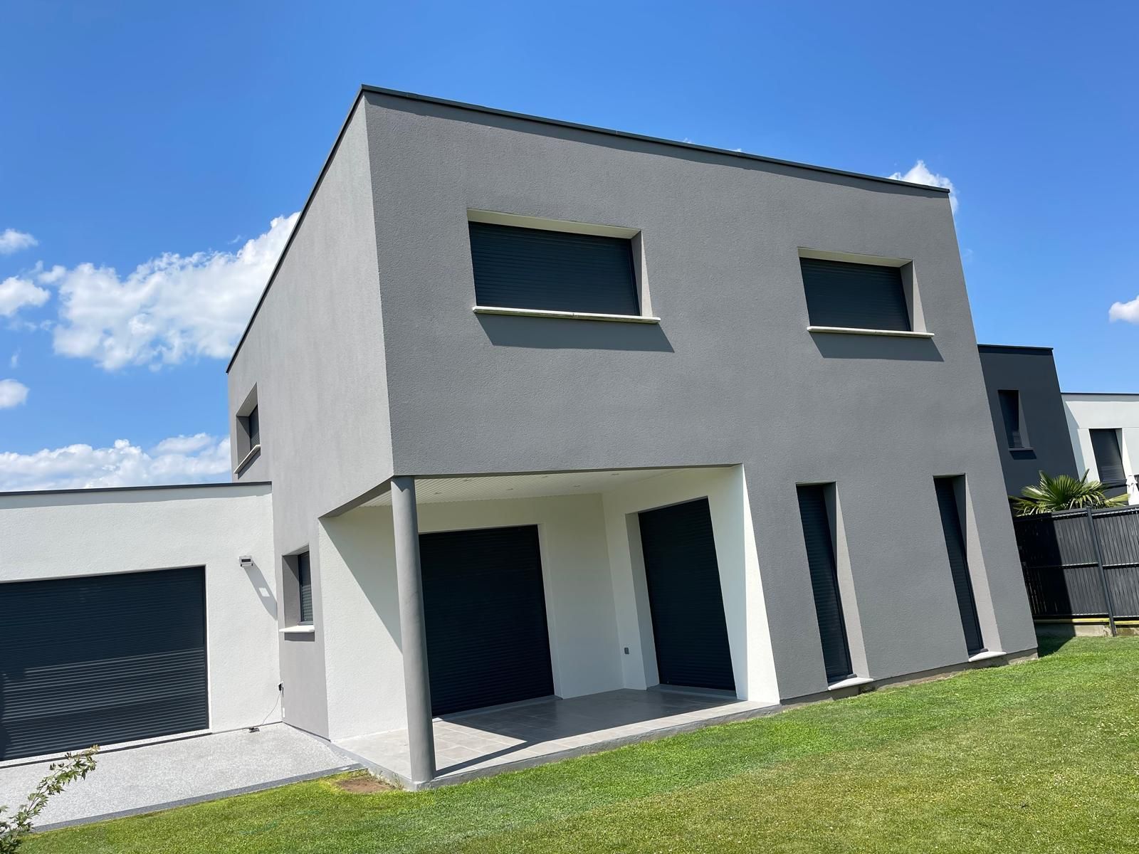 Maison moderne grise de deux étages avec porte de garage et fenêtres noires, située sur une colline verdoyante sous un ciel bleu.