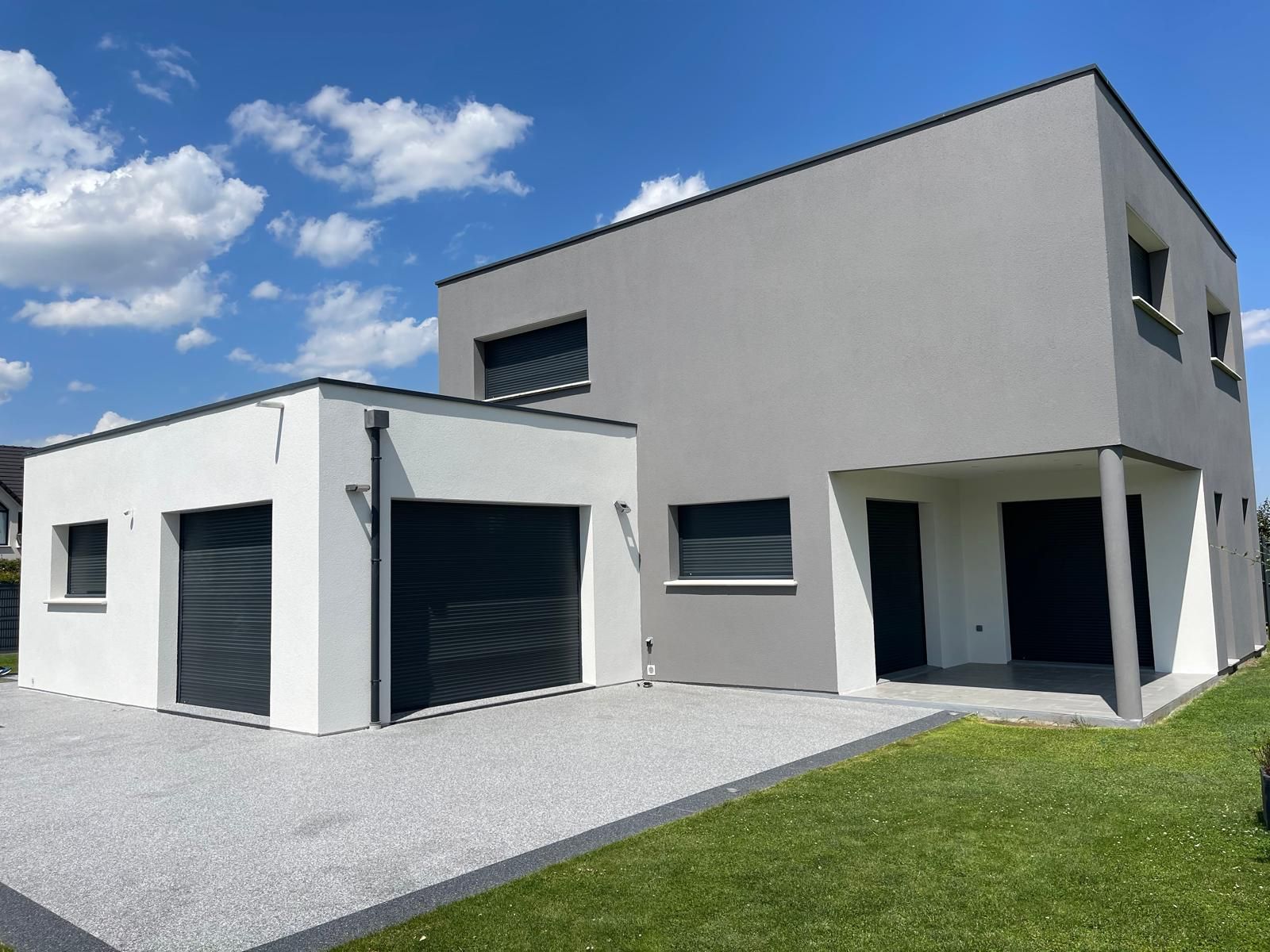 Maison moderne à deux étages avec façade grise et blanche, portes de garage noires et allée de gravier, par une journée ensoleillée.