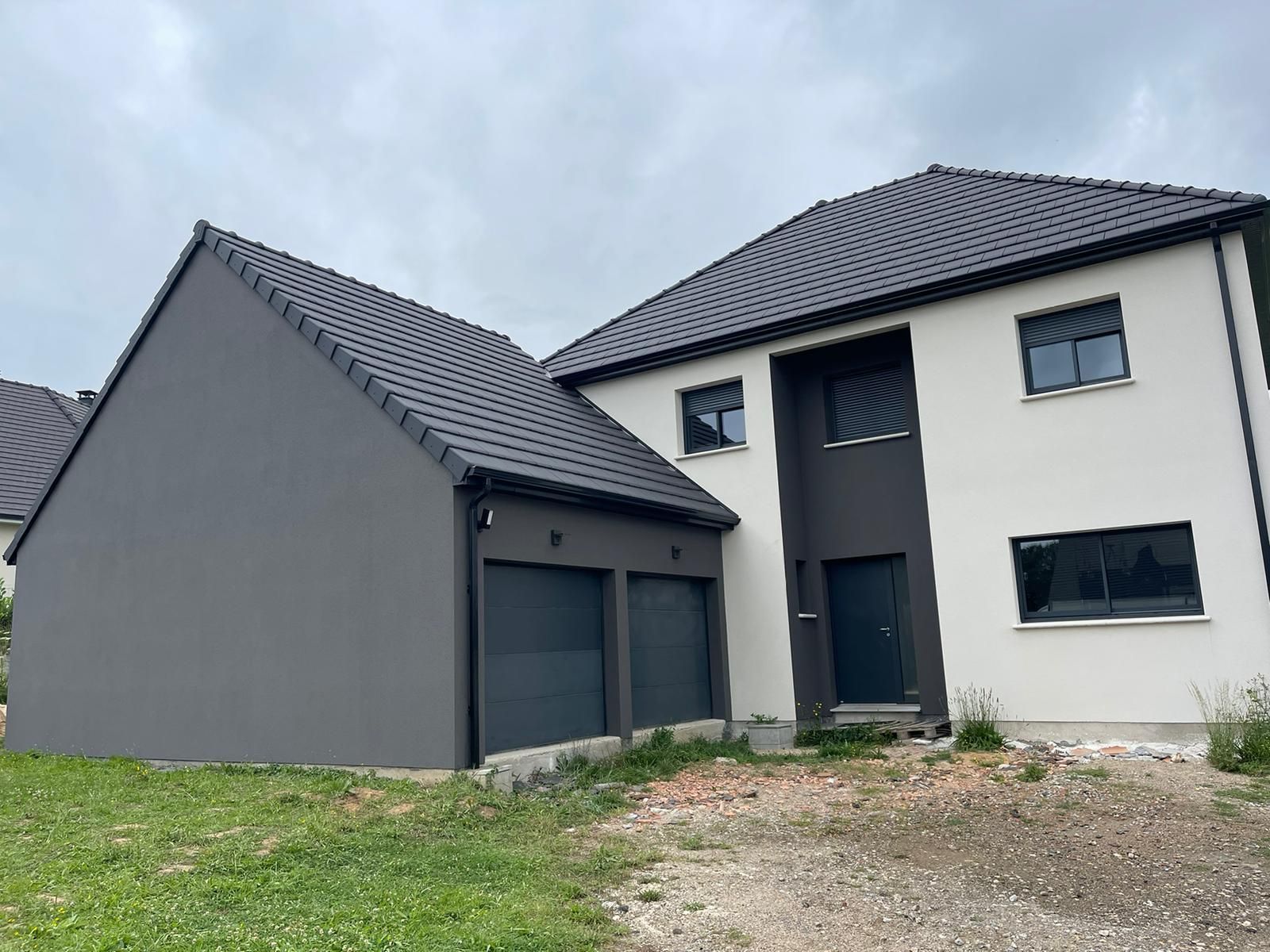 Maison moderne à deux étages avec garage et toit gris. Façade blanche, touches de gris foncé, fenêtres. Ciel nuageux, jardin gazonné.