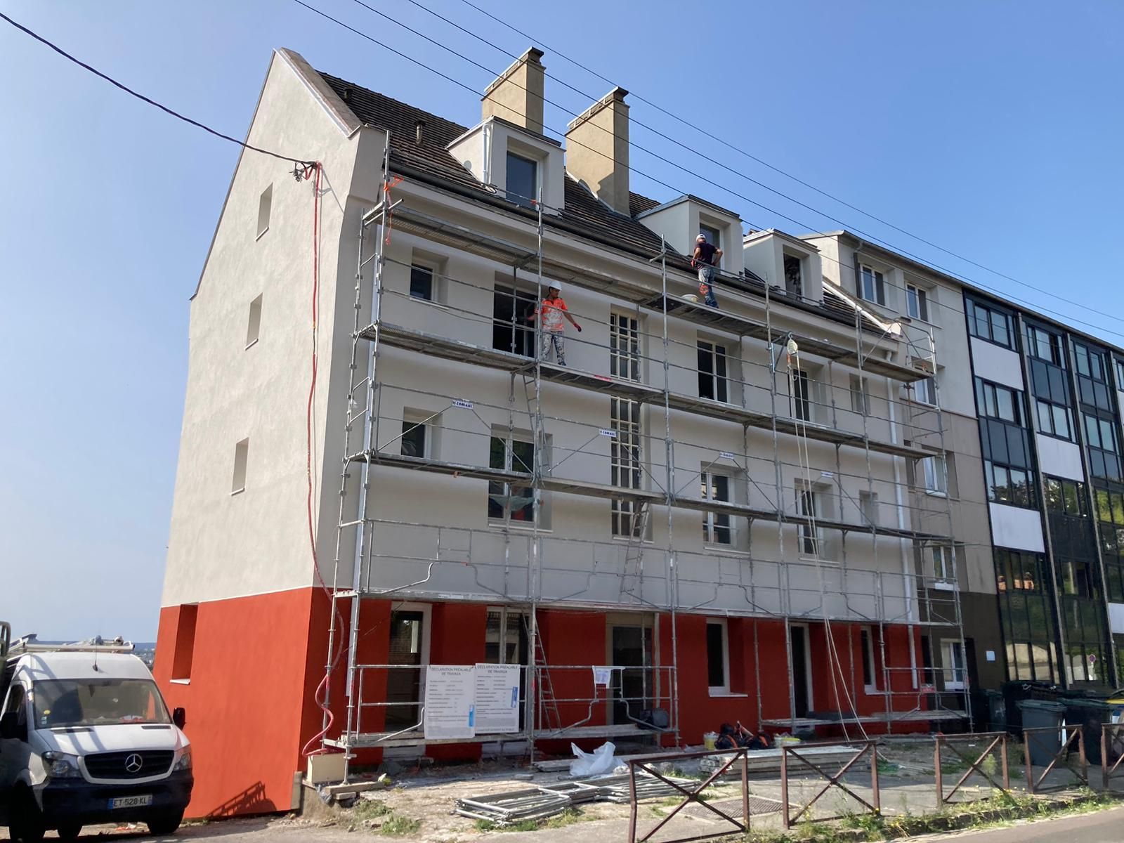 Bâtiment en rénovation, avec échafaudages et ouvriers. Façade rouge et blanche, ciel bleu.