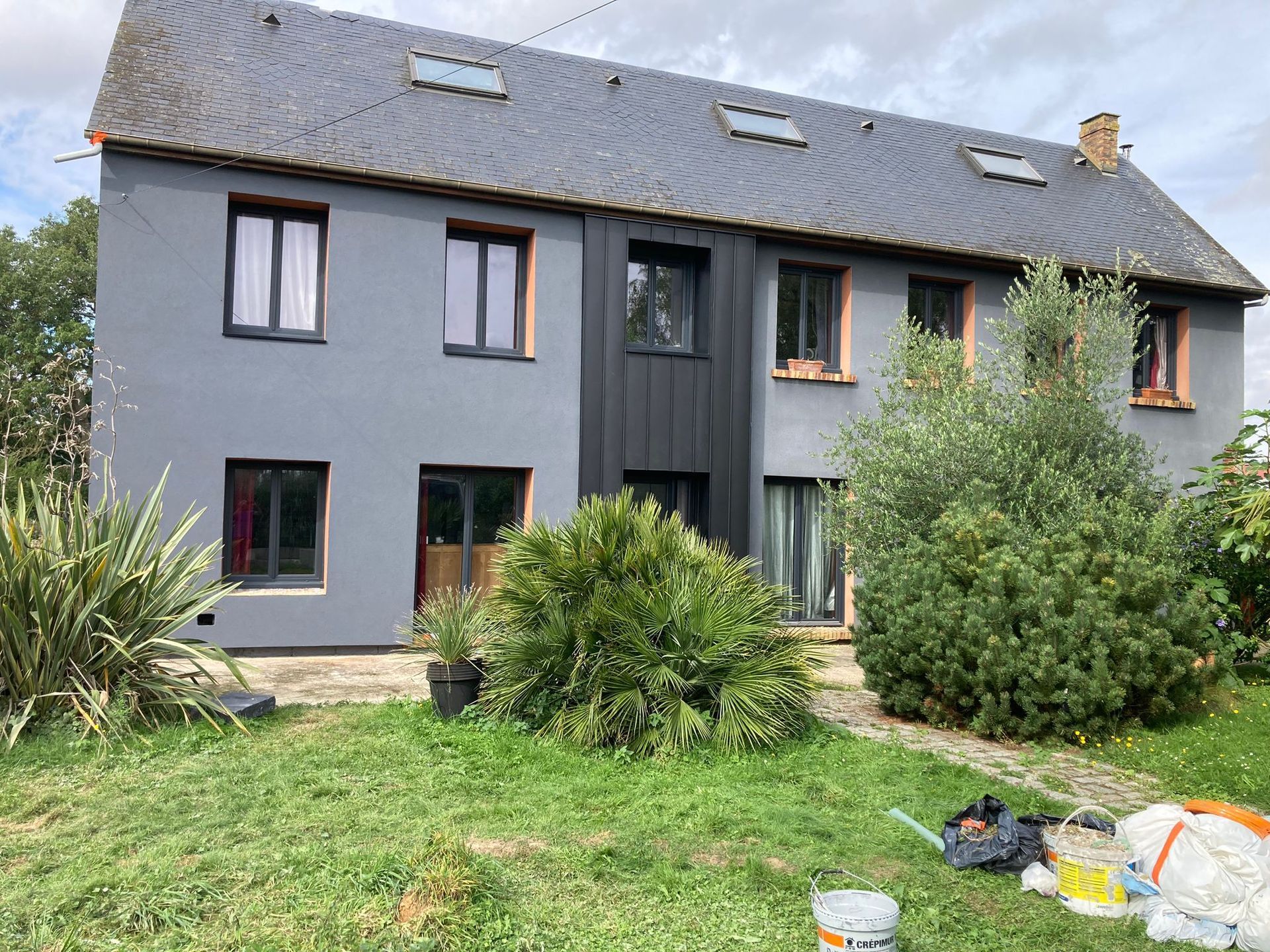 Maison grise aux fenêtres sombres, toit en ardoise et jardin gazonné.