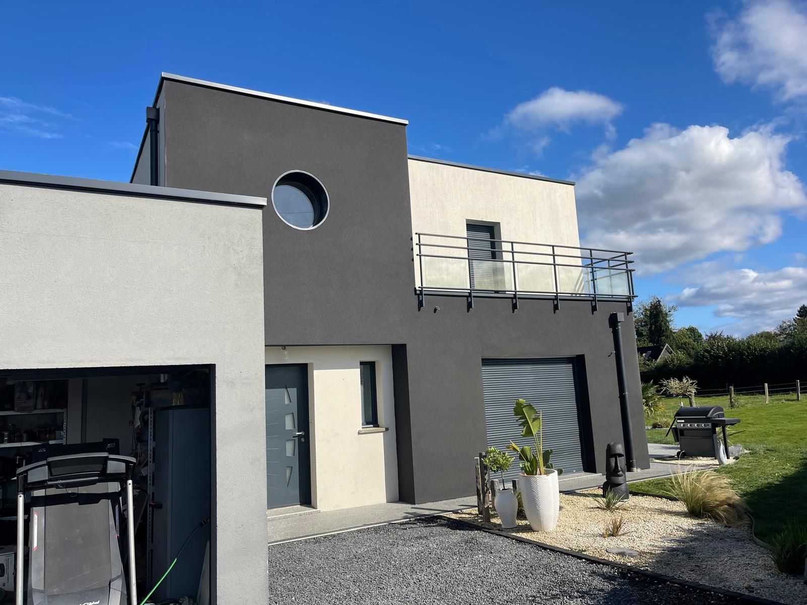 Maison moderne à deux étages avec des murs gris foncé et beige clair, un balcon et une fenêtre ronde sous un ciel bleu.