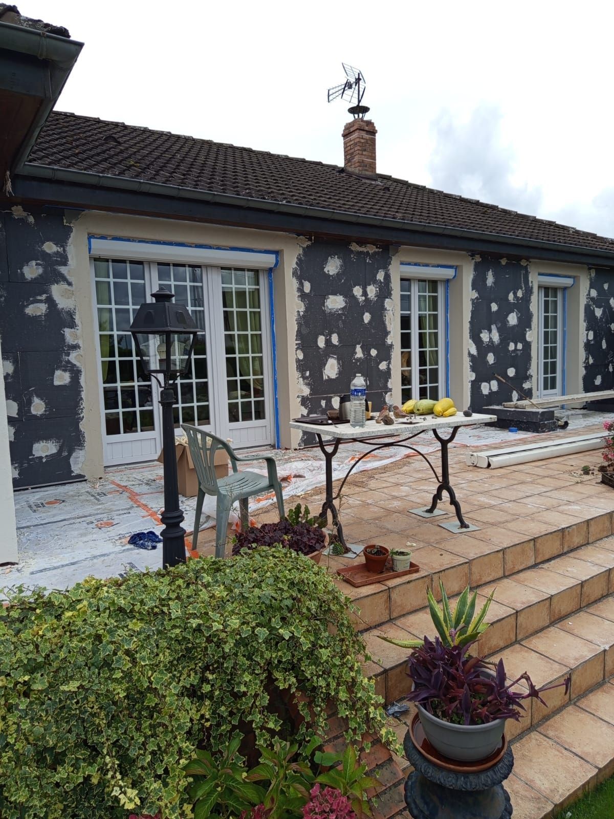 Extérieur d'une maison en rénovation. Des plaques d'enduit gris recouvrent la façade ; patio avec mobilier et plantes.