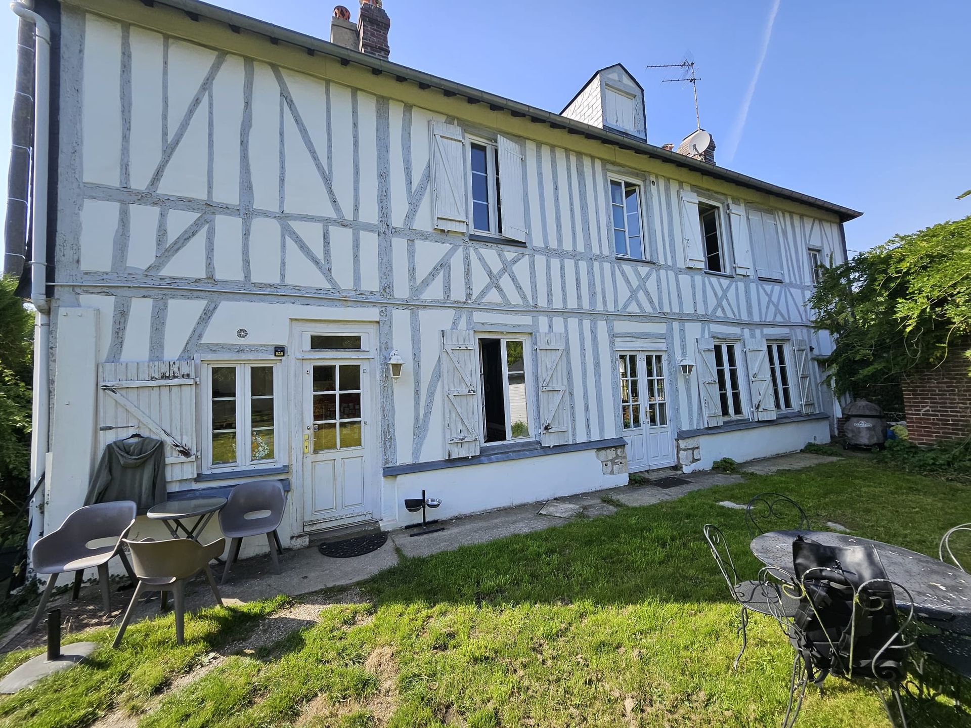 Bâtiment à ossature en bois blanche de deux étages avec volets ouverts, petit patio avec chaises sur une colline herbeuse.