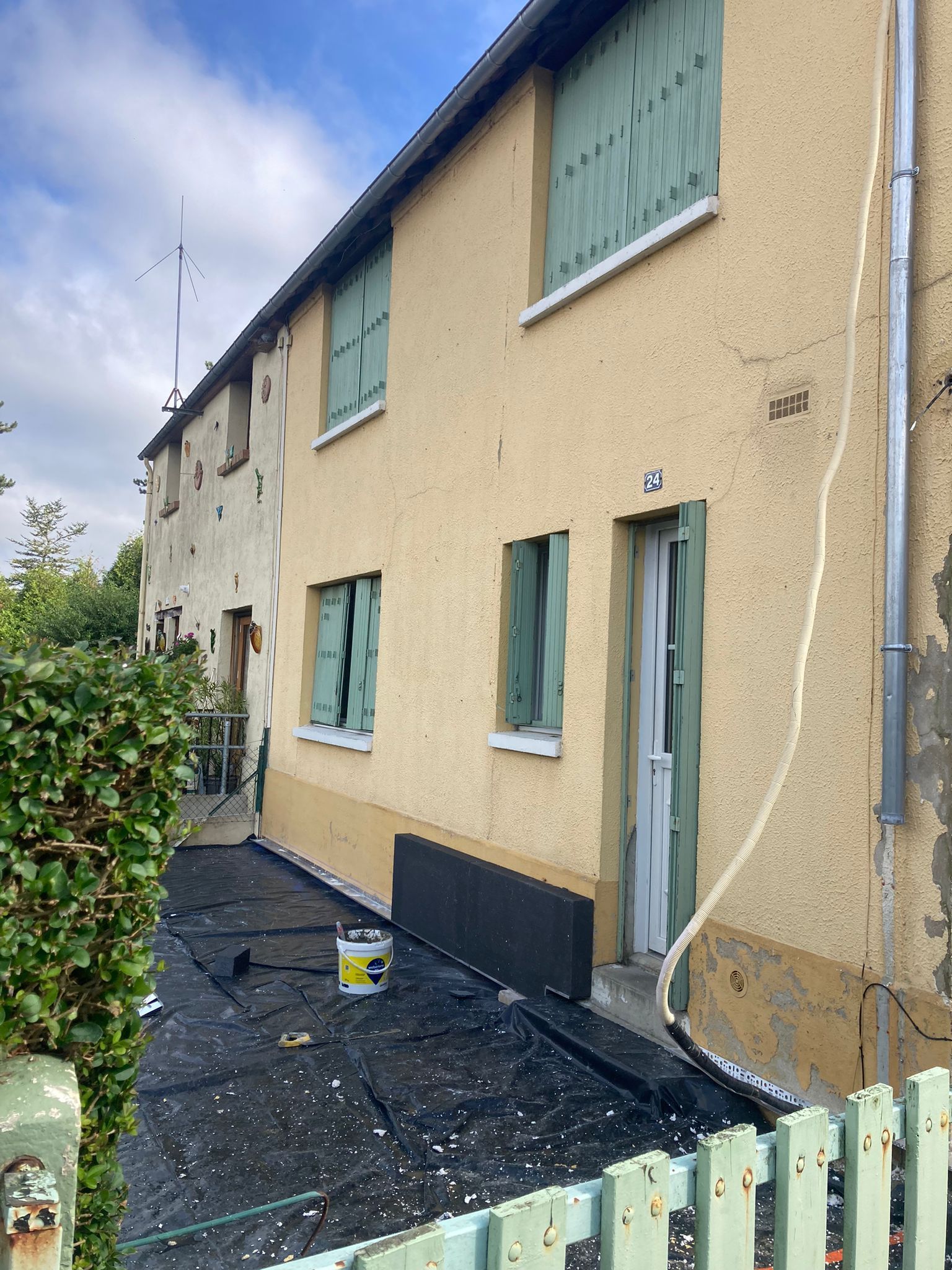 Maison jaune aux volets verts, en cours de rénovation. Matériau noir au sol, un seau et une clôture.
