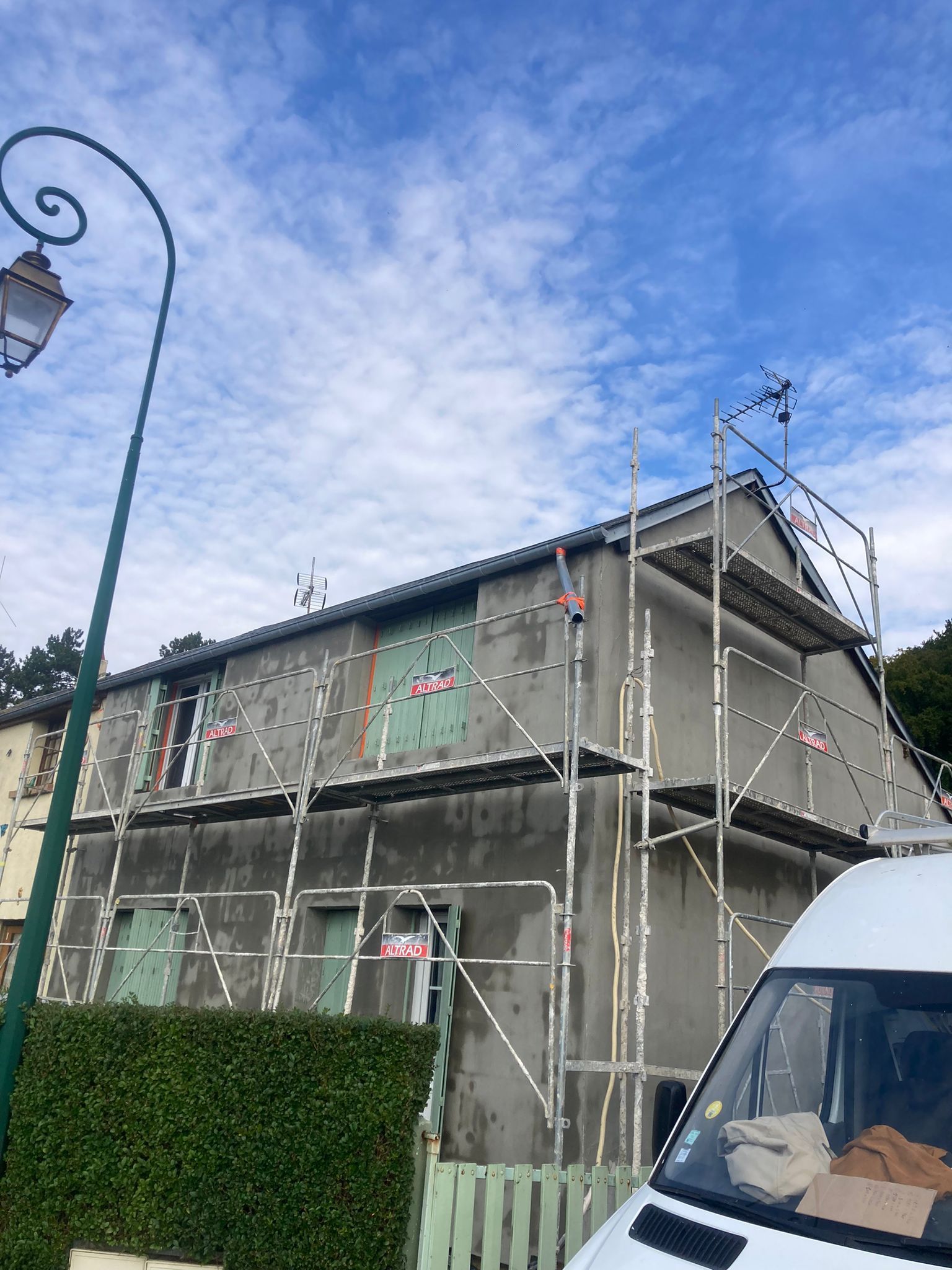 Une maison en rénovation avec des échafaudages ; murs gris, volets verts et ciel bleu.