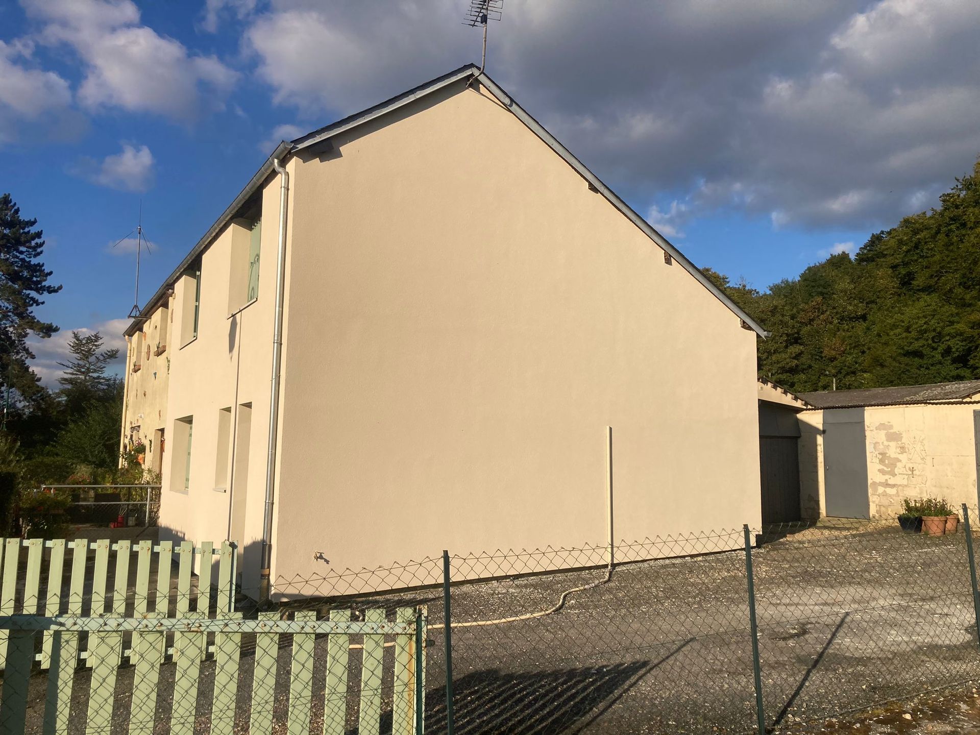 Bâtiment beige de deux étages avec un mur lisse, jouxtant un abri de jardin et une clôture verte sous un ciel partiellement nuageux.