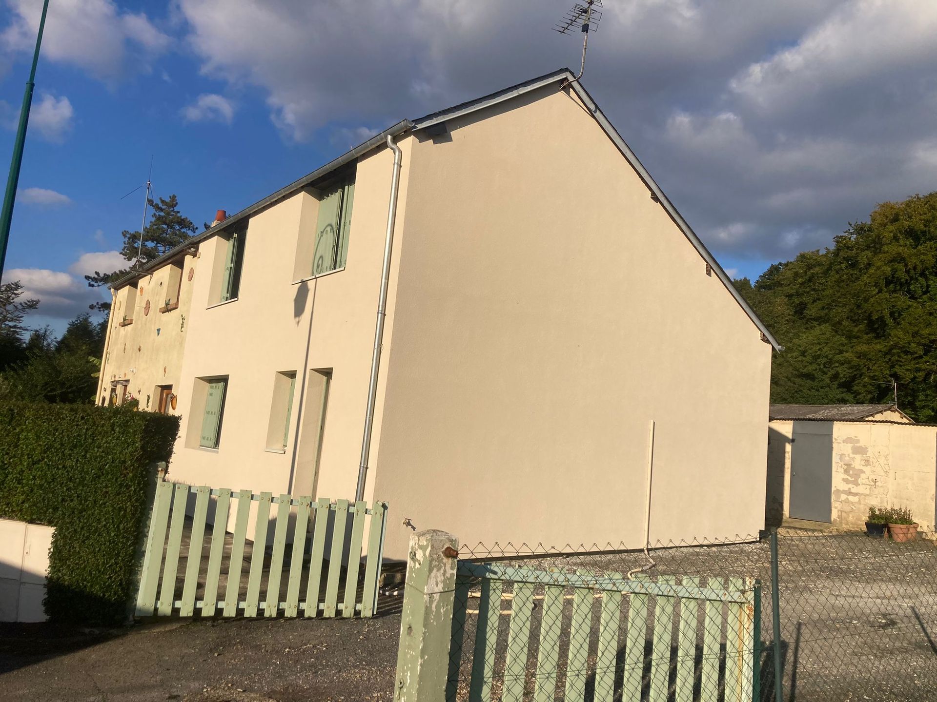 Maison beige avec des volets vert pâle et une clôture blanche. Ciel bleu et quelques arbres.