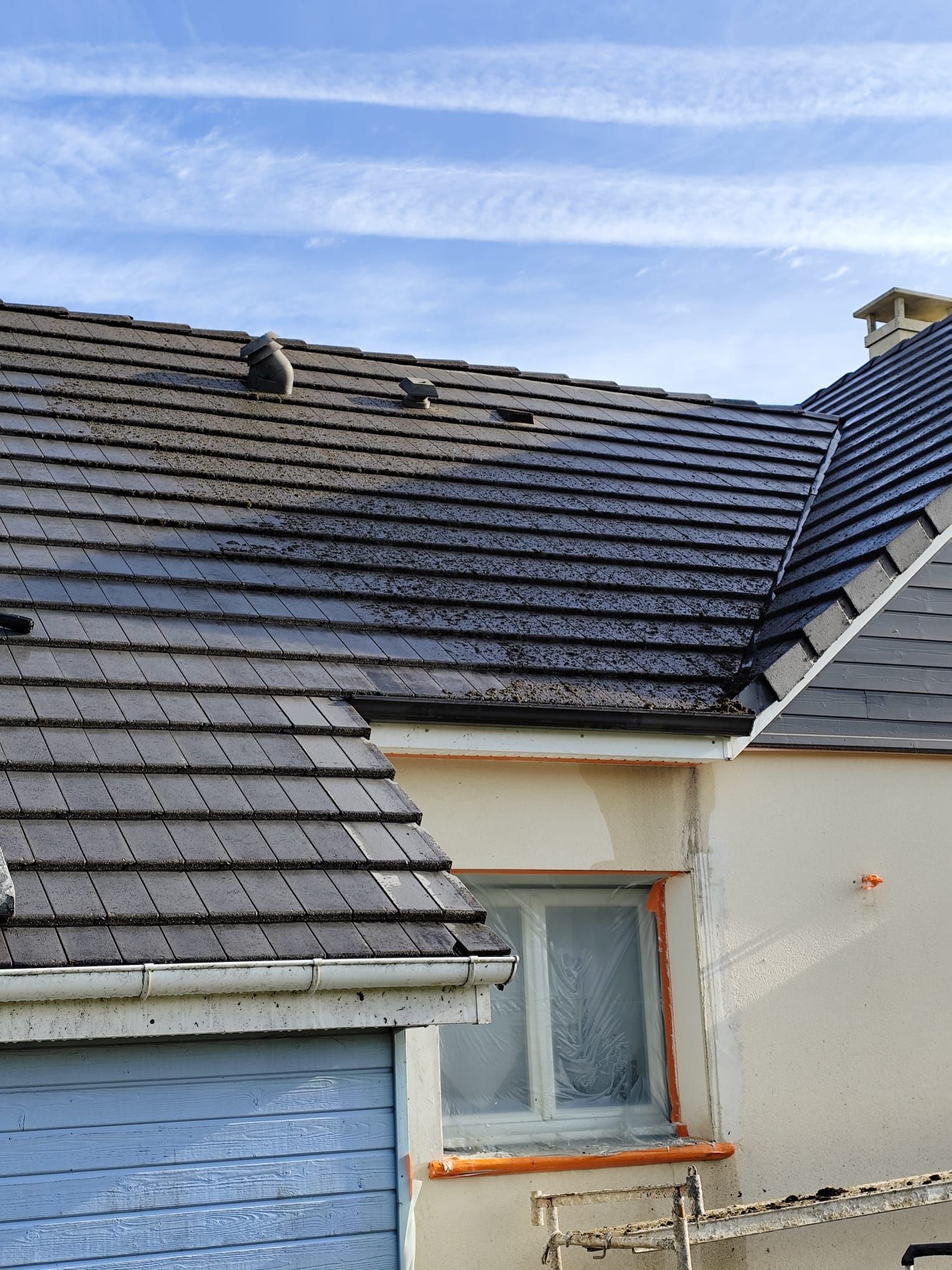 Toiture en tuiles sombres sur une maison sous un ciel bleu partiellement nuageux, avec une fenêtre et une porte de garage bleue visibles.