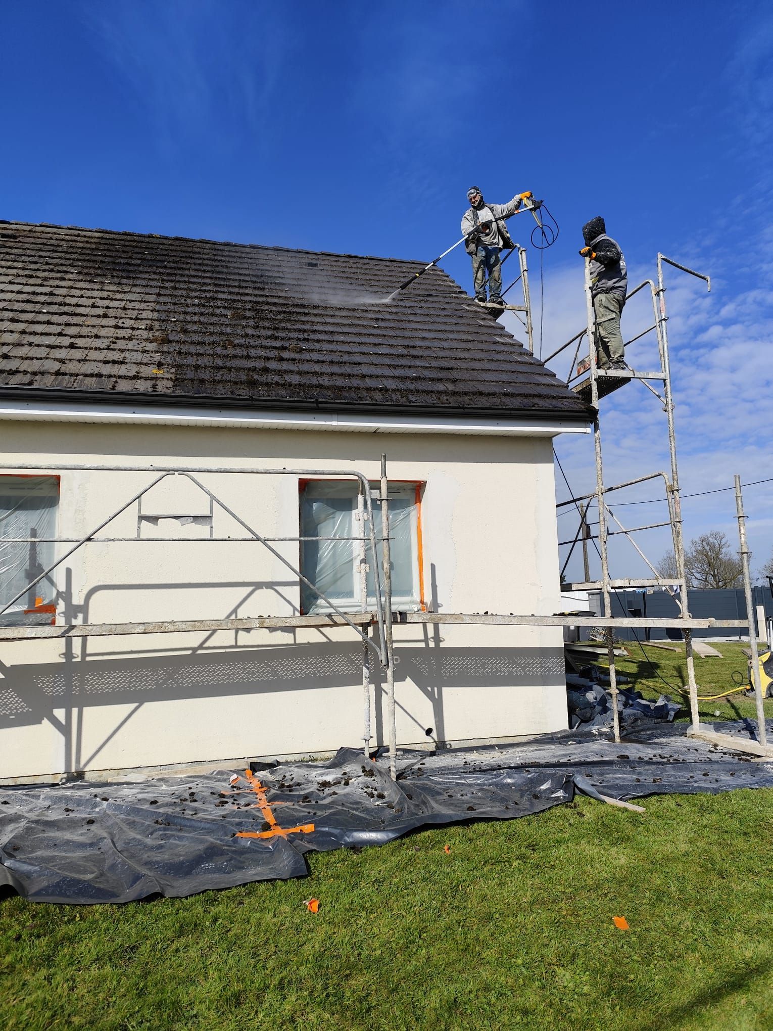 Des ouvriers sur des échafaudages pulvérisent un produit nettoyant sur un toit, avec une maison et un ciel bleu en arrière-plan.