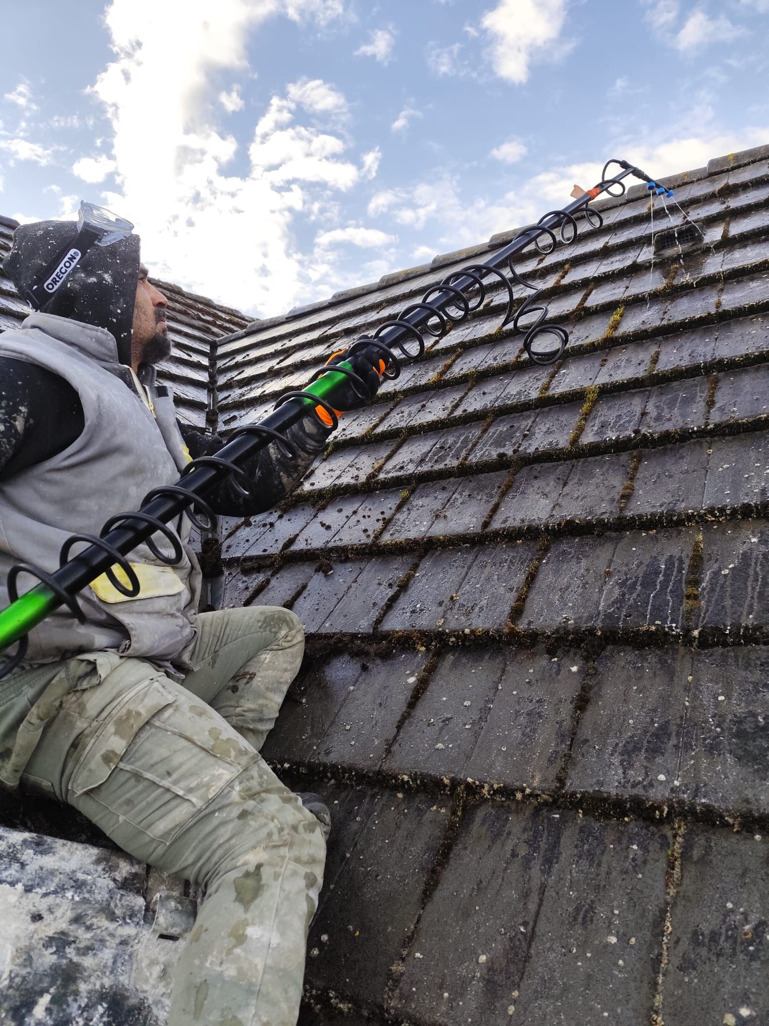 Une personne nettoie un toit avec un outil à long manche. Le ciel est nuageux. Le toit est recouvert de mousse.