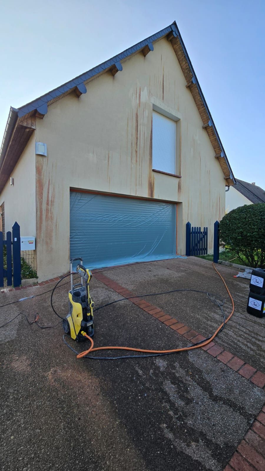 Nettoyage haute pression de la façade du bâtiment ; porte de garage recouverte, nettoyeur haute pression sur l'allée.