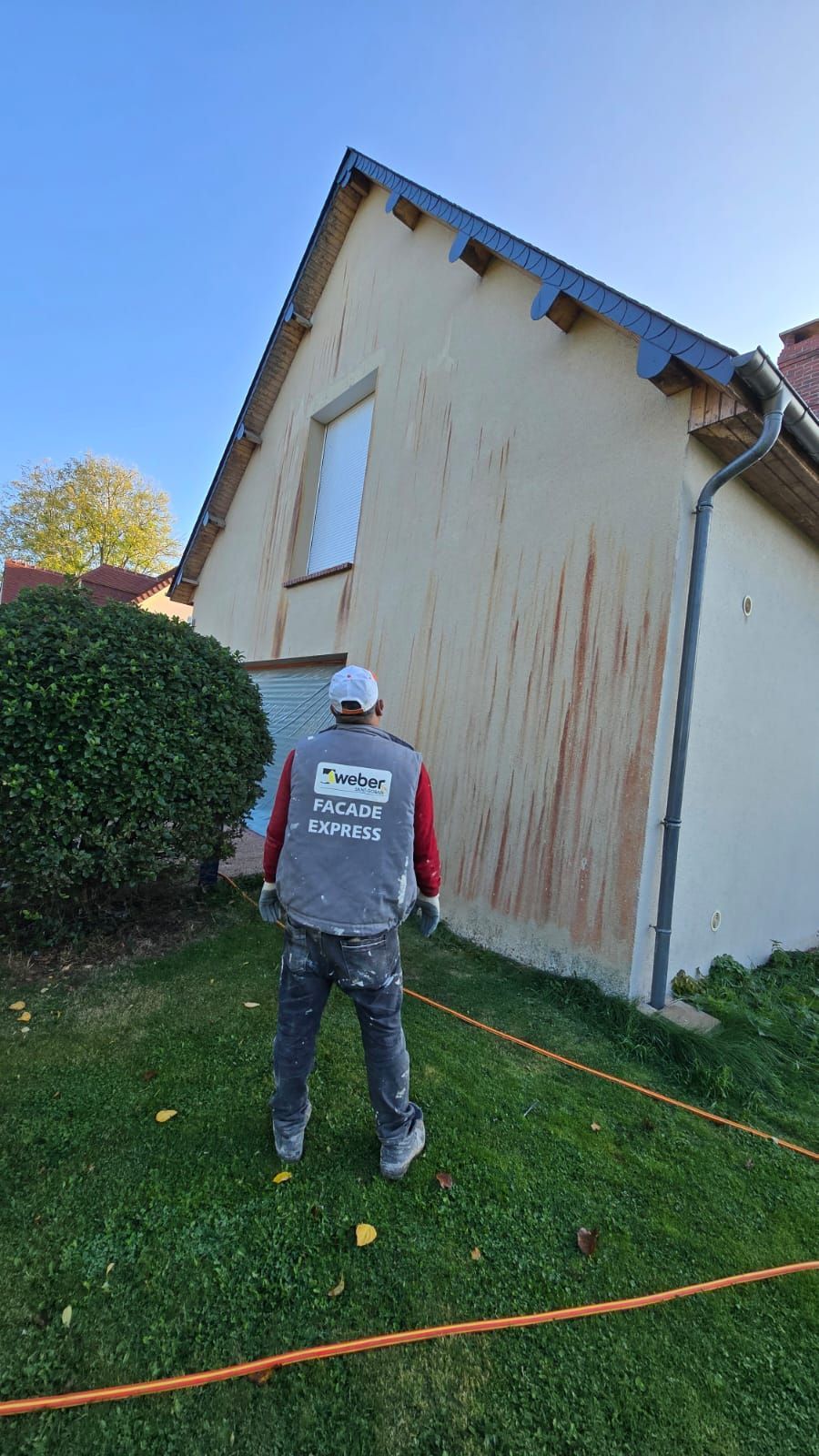 Une personne en tenue de travail examine une maison présentant des taches verticales ; pelouse verte, ciel bleu.