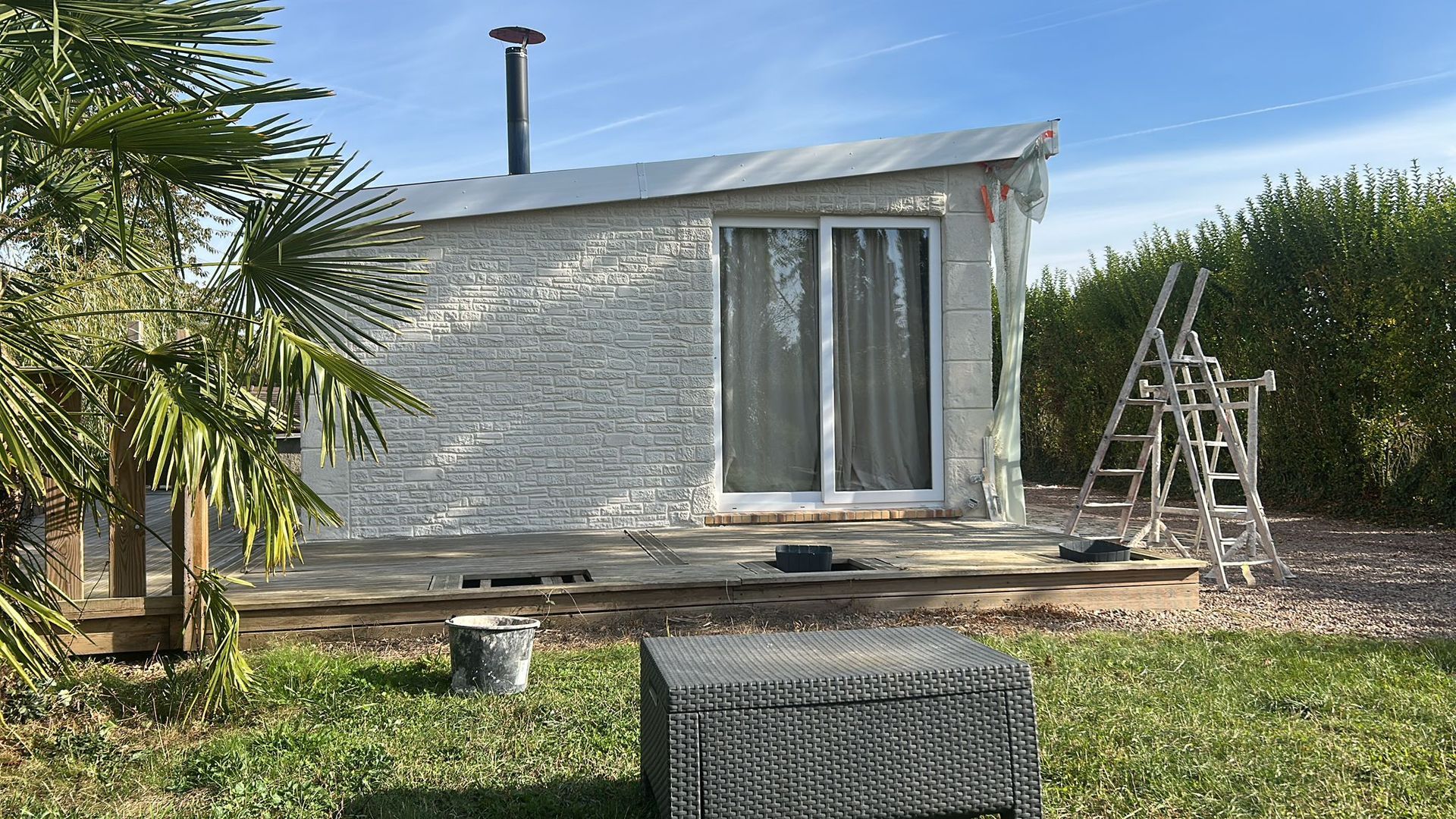 Petit cabanon gris, inachevé, avec portes coulissantes en verre, terrasse et échelle à l'extérieur, par une journée ensoleillée.