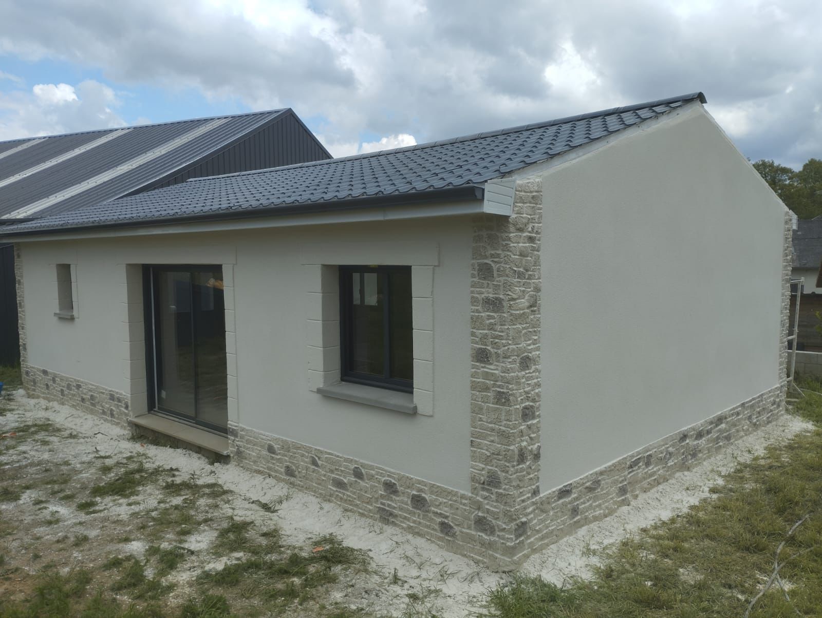 Petite maison aux murs en stuc clair, avec des touches de pierre, un toit et des encadrements de fenêtres noirs.