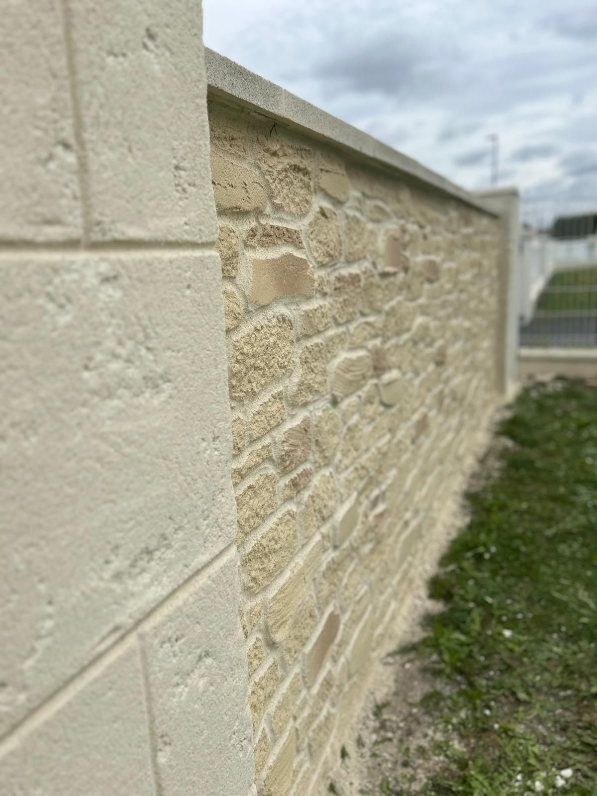 Mur en pierre à la surface texturée beige, chapiteau et poteaux d'angle. Herbe verte au pied ; ciel couvert.