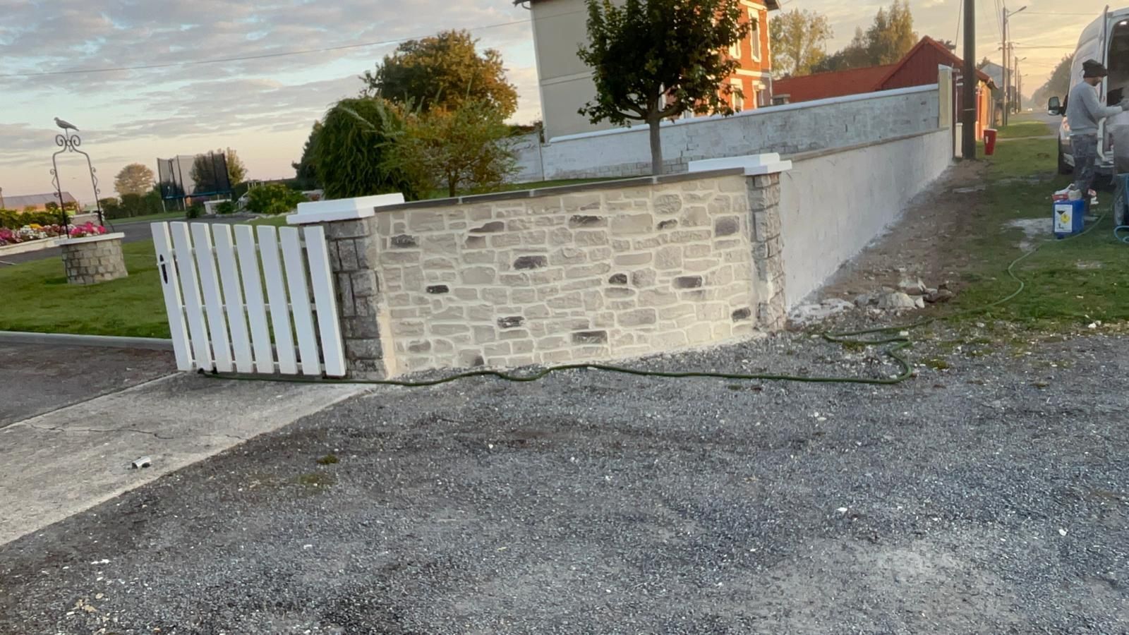 Un muret en pierre avec une clôture blanche et un enduit, bordant une route ; une maison et des arbres en arrière-plan.