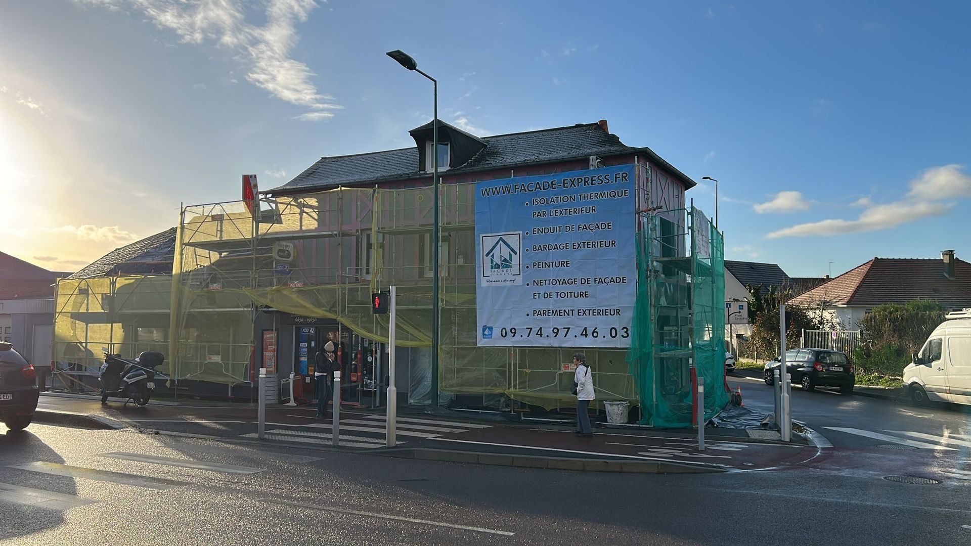 Bâtiment en rénovation, recouvert d'échafaudages et de filets de sécurité. Panneau de chantier visible. Cadre extérieur ensoleillé.