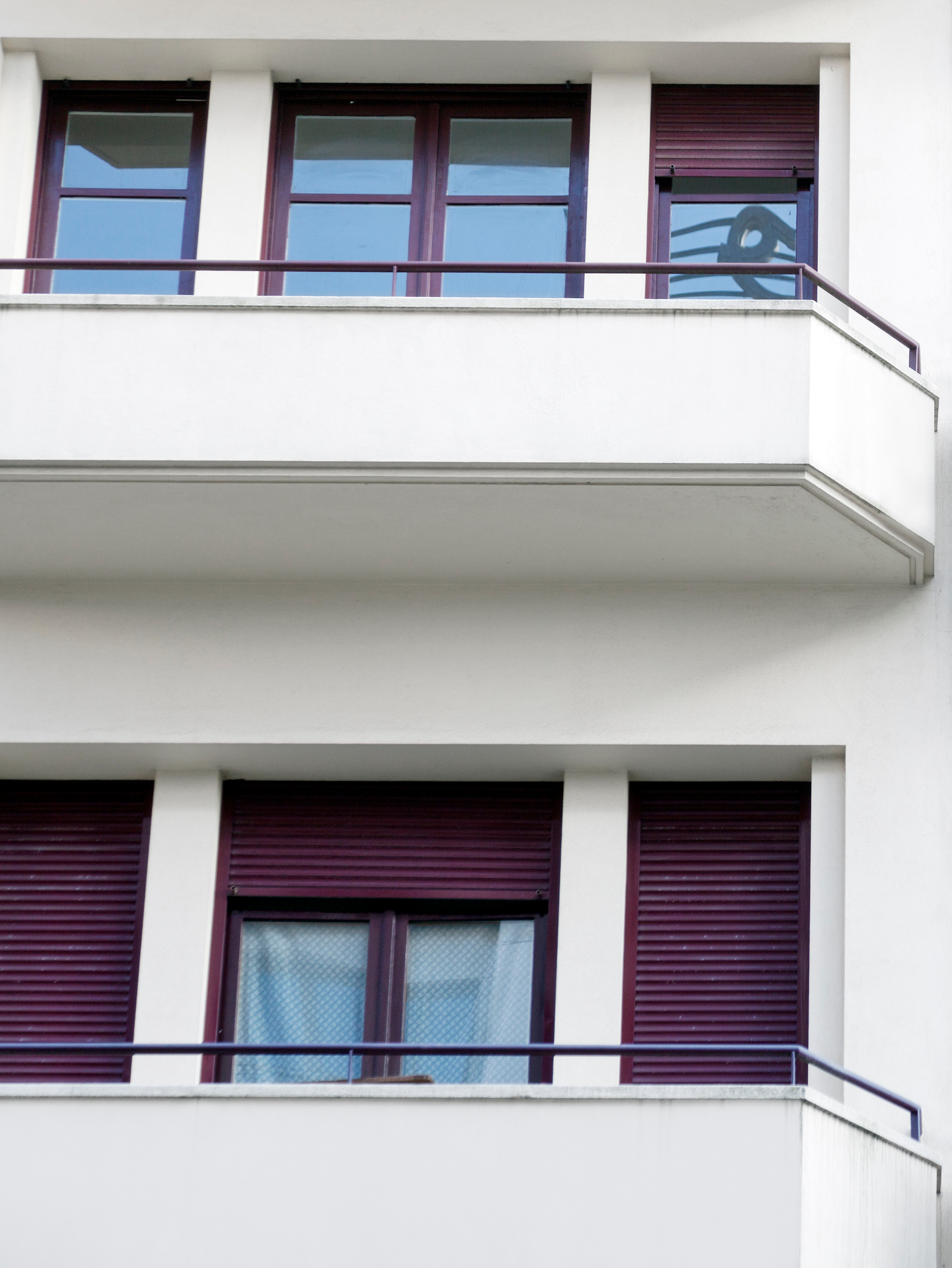 Immeuble blanc aux encadrements de fenêtres et volets bordeaux. Balcons à balustrades bleues.