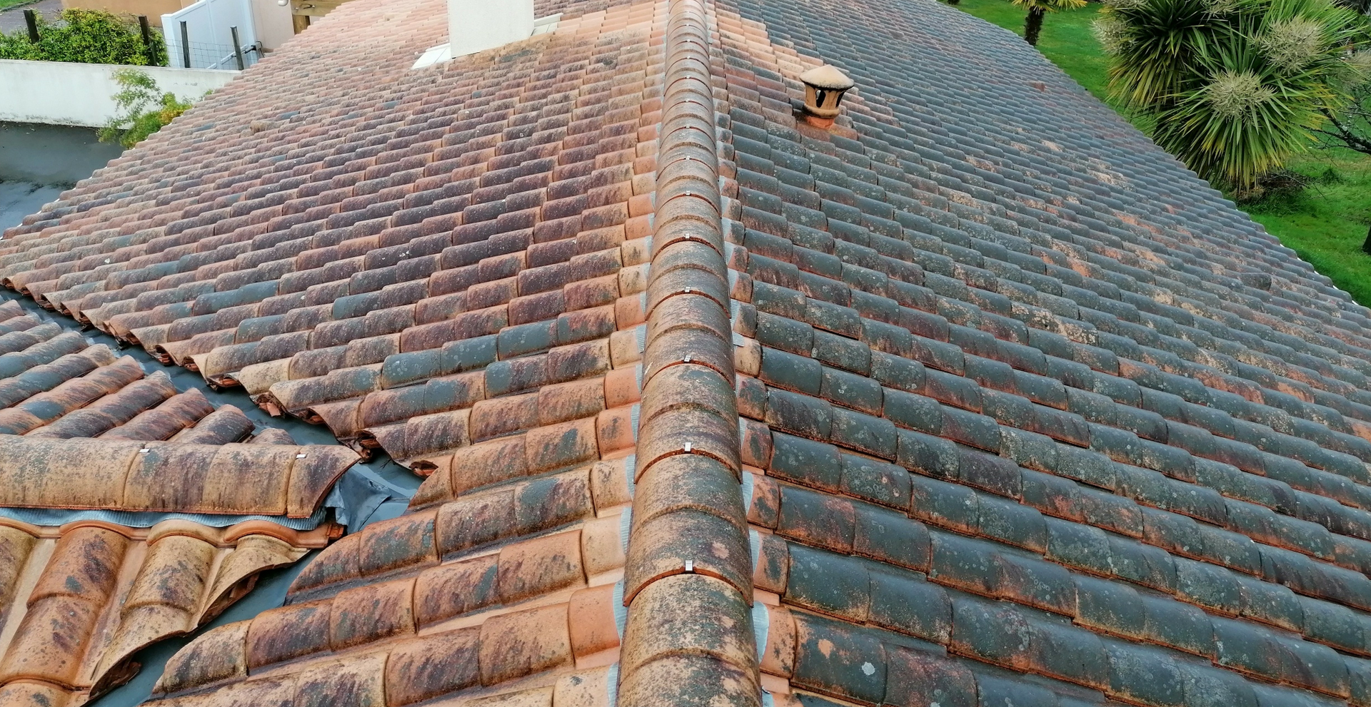 Toiture en tuiles brunes, montrant des signes d'usure, avec une cheminée et de la végétation verte en arrière-plan.