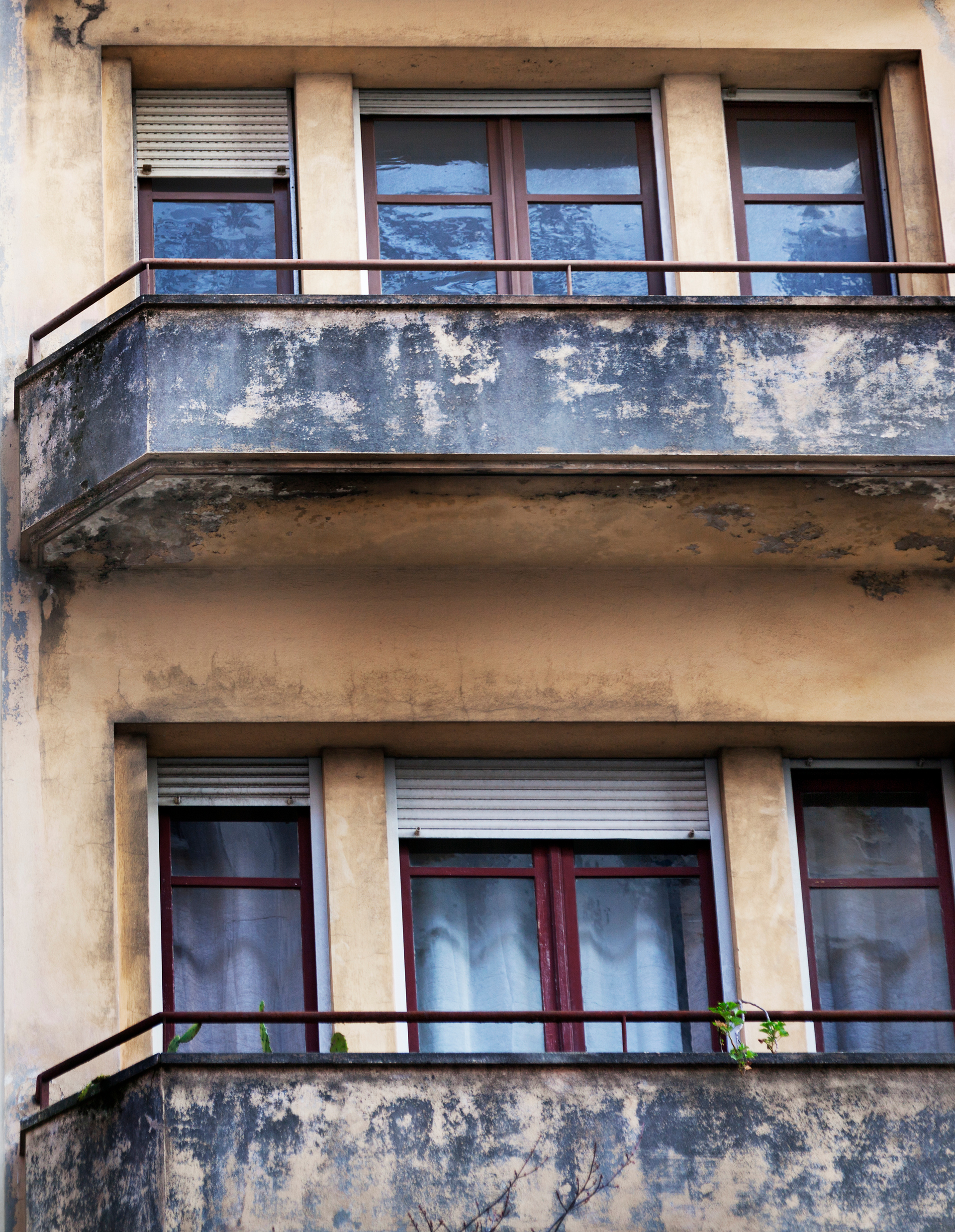 Extérieur d'un bâtiment délabré avec balcons et fenêtres, encadrements bruns, peinture défraîchie, murs beige clair et quelques plantes.