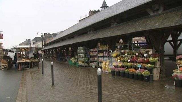 Buchy - Marché en plein air sous un auvent en bois ; des étals de fleurs et de produits frais bordent l'allée.