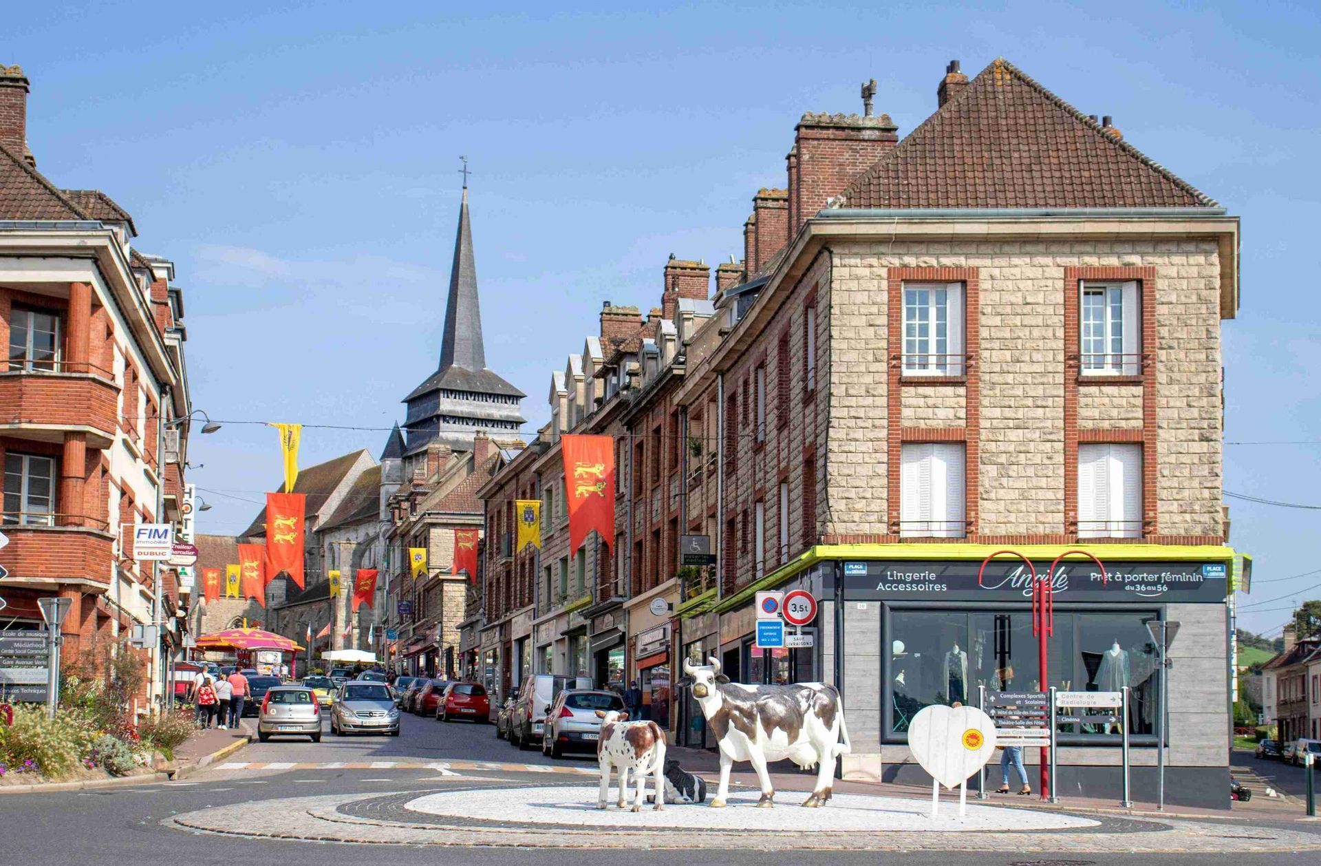 Neufchâtel-en-Bray - Rue d'une ville normande avec rond-point pavé, vaches, magasins et clocher d'église.
