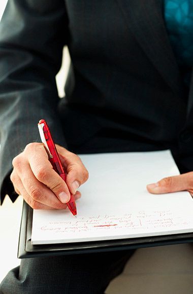 Una persona vestida con un traje oscuro sostiene un bolígrafo rojo y escribe en un bloc de notas blanco sujeto a un portapapeles.