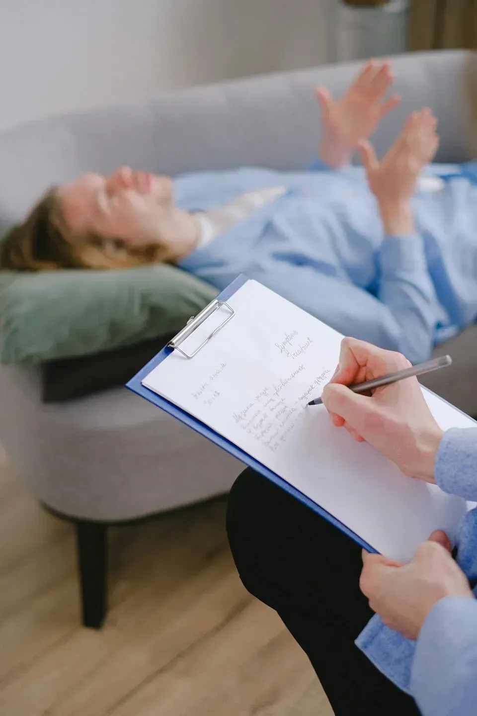 Un profesional toma notas en un portapapeles mientras una persona yace en un sofá al fondo, gesticulando durante una sesión de terapia.