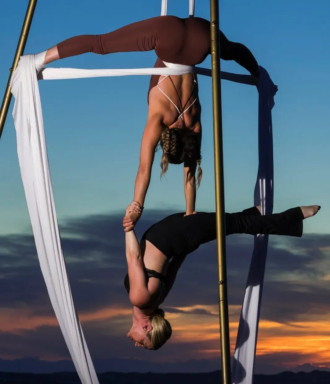 Dos artistas aéreos posan boca abajo en un aro de tela al atardecer, uno sujetando la mano del otro.