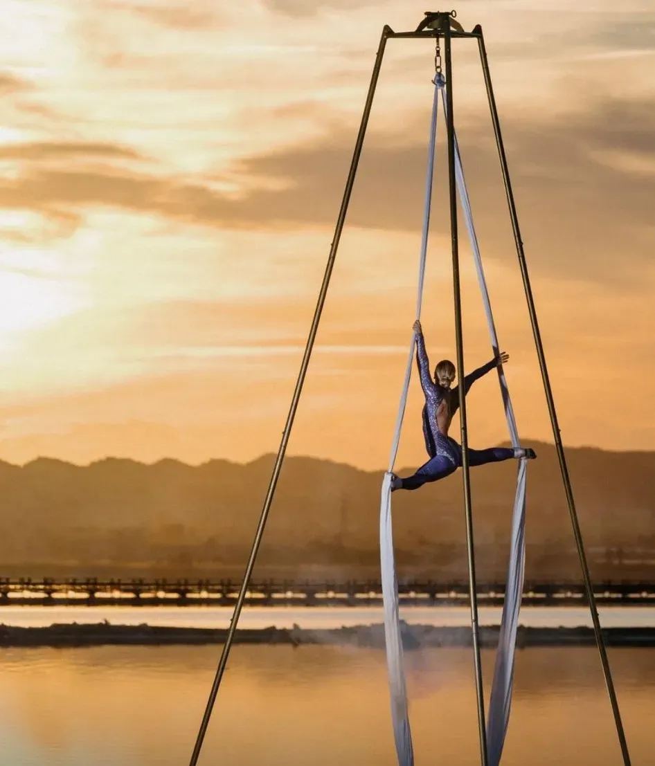 Acróbata aéreo suspendido sobre el agua al atardecer, enmarcado por cuerdas colgantes contra un cielo cálido.